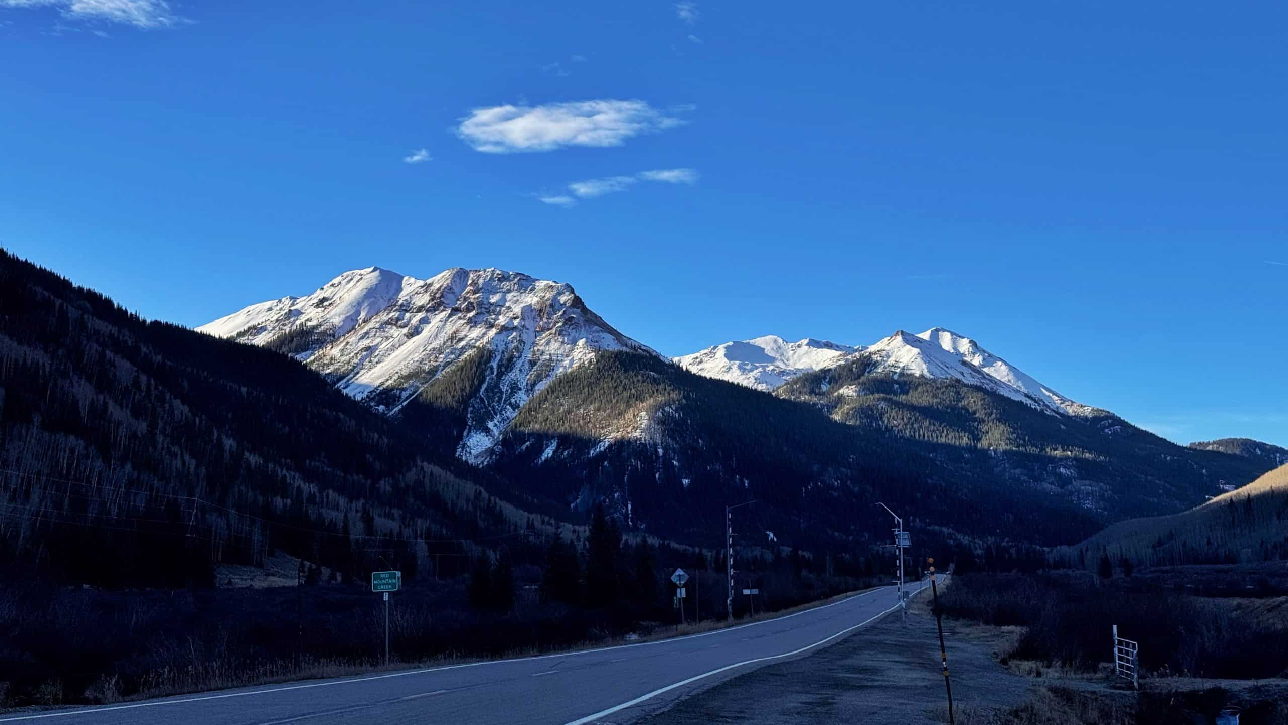 A scenic view of the Million-Dollar Highway featuring a winding road through a mountain valley with snow-capped peaks and evergreen forests under a clear blue sky, 