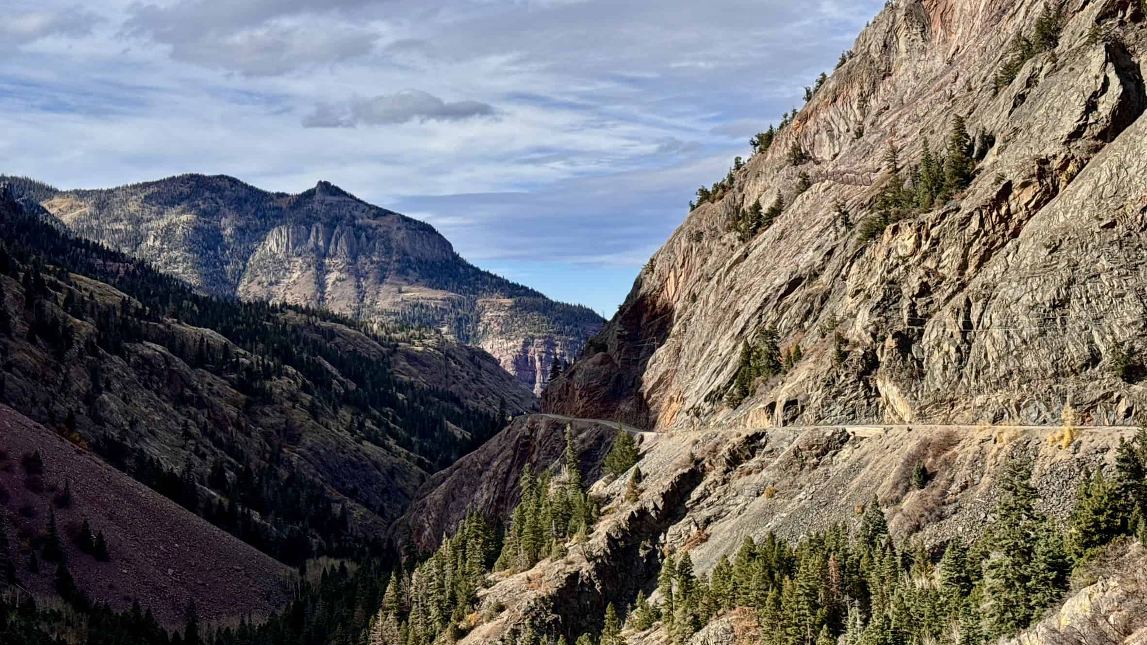 A scenic road near Ouray, carved into the side of a steep mountain surrounded with dramatic cliffs and forested slopes under a partly cloudy sky
