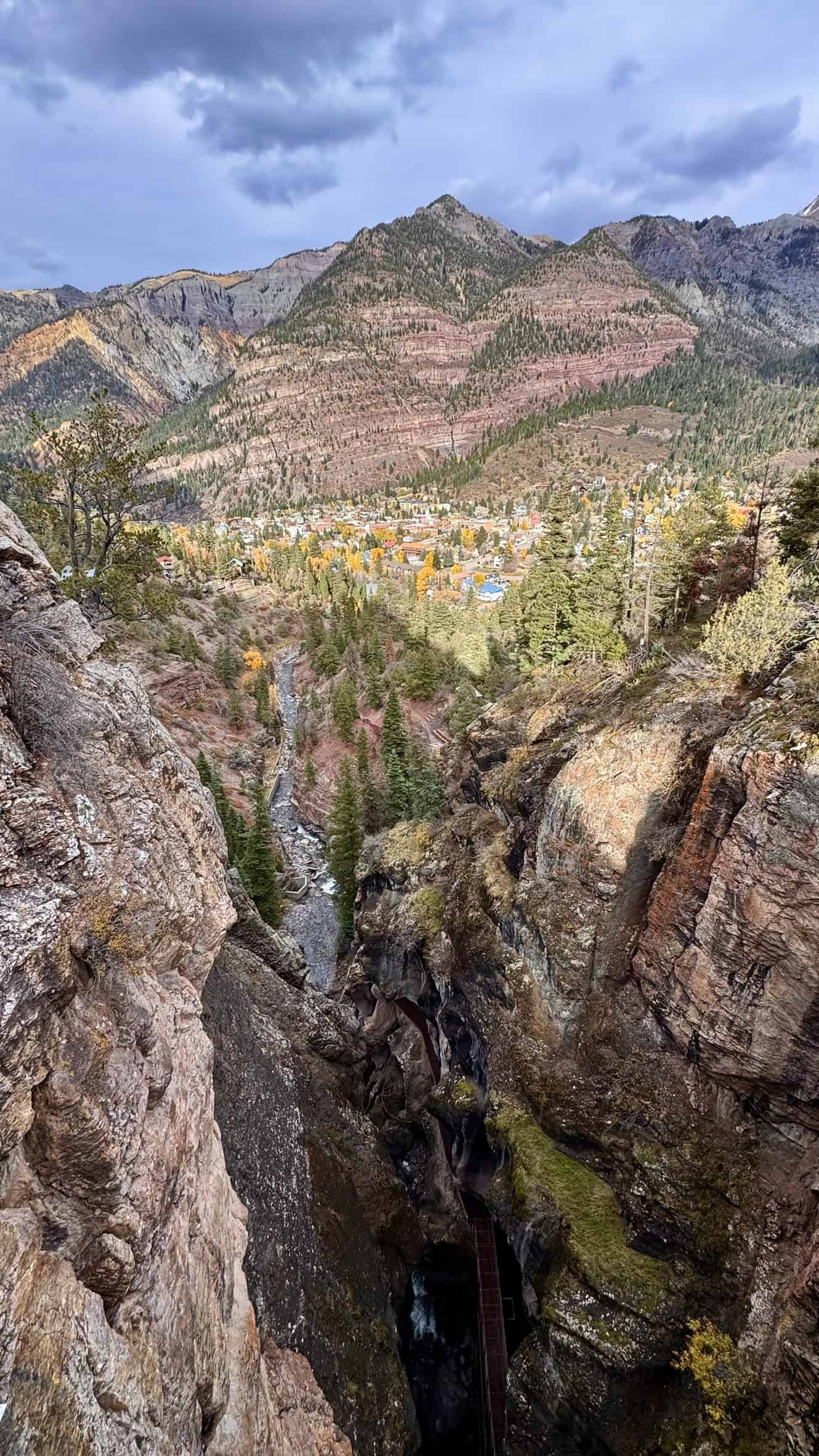 A sweeping view from the High Bridge Trail overlooking a gorge, and a mountain valley with Ouray nestled among autumn-colored trees under a cloudy sky.