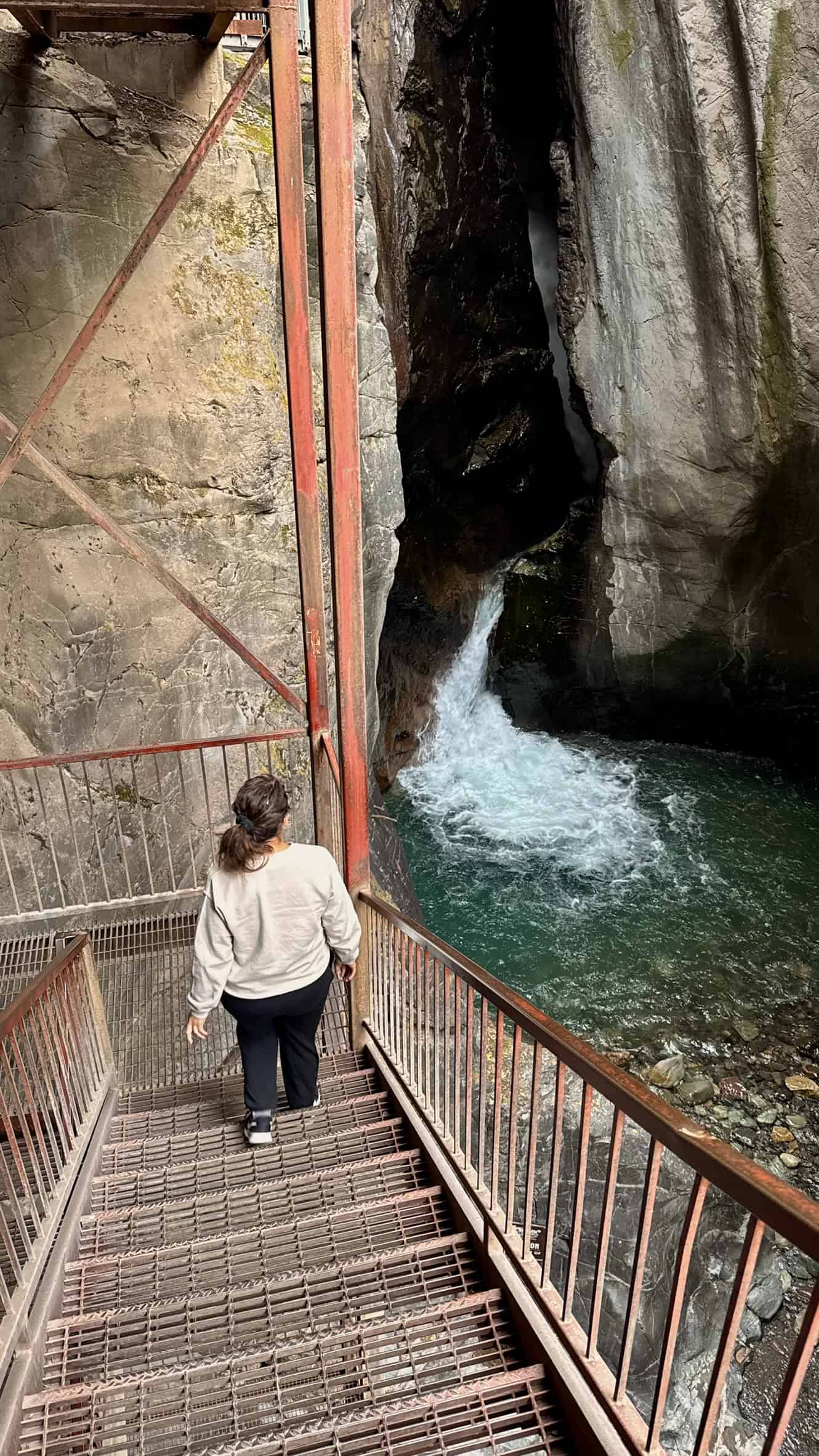 A woman walks down metal stairs in a deep canyon gorge looking towards a waterfall flowing into a emerald green pool, highlighting one of the best things to do in Ouray. 
