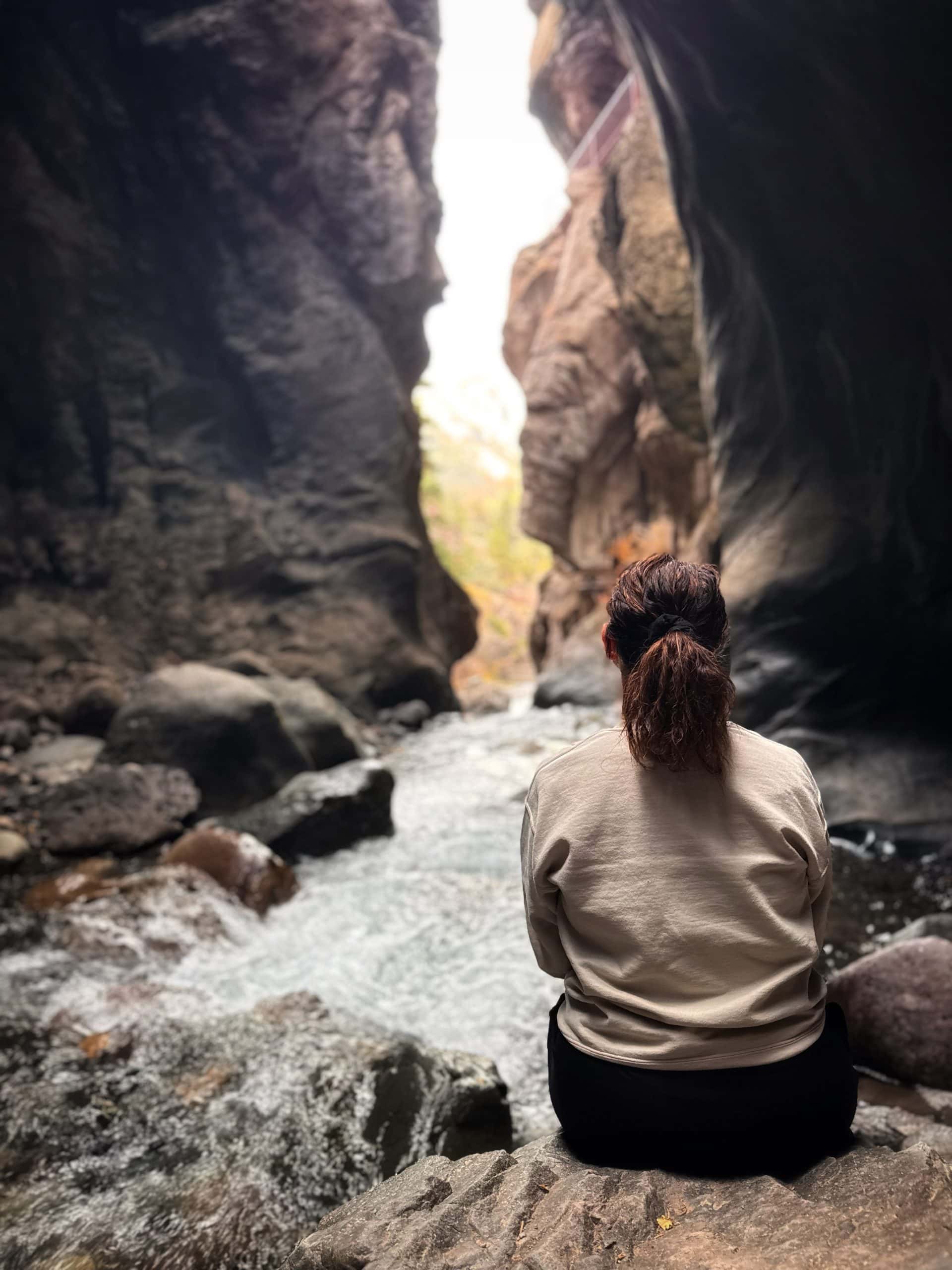 A woman sits on a rock overlooking a rushing river in a narrow gorge, enjoying the serene natural beauty of Ouray