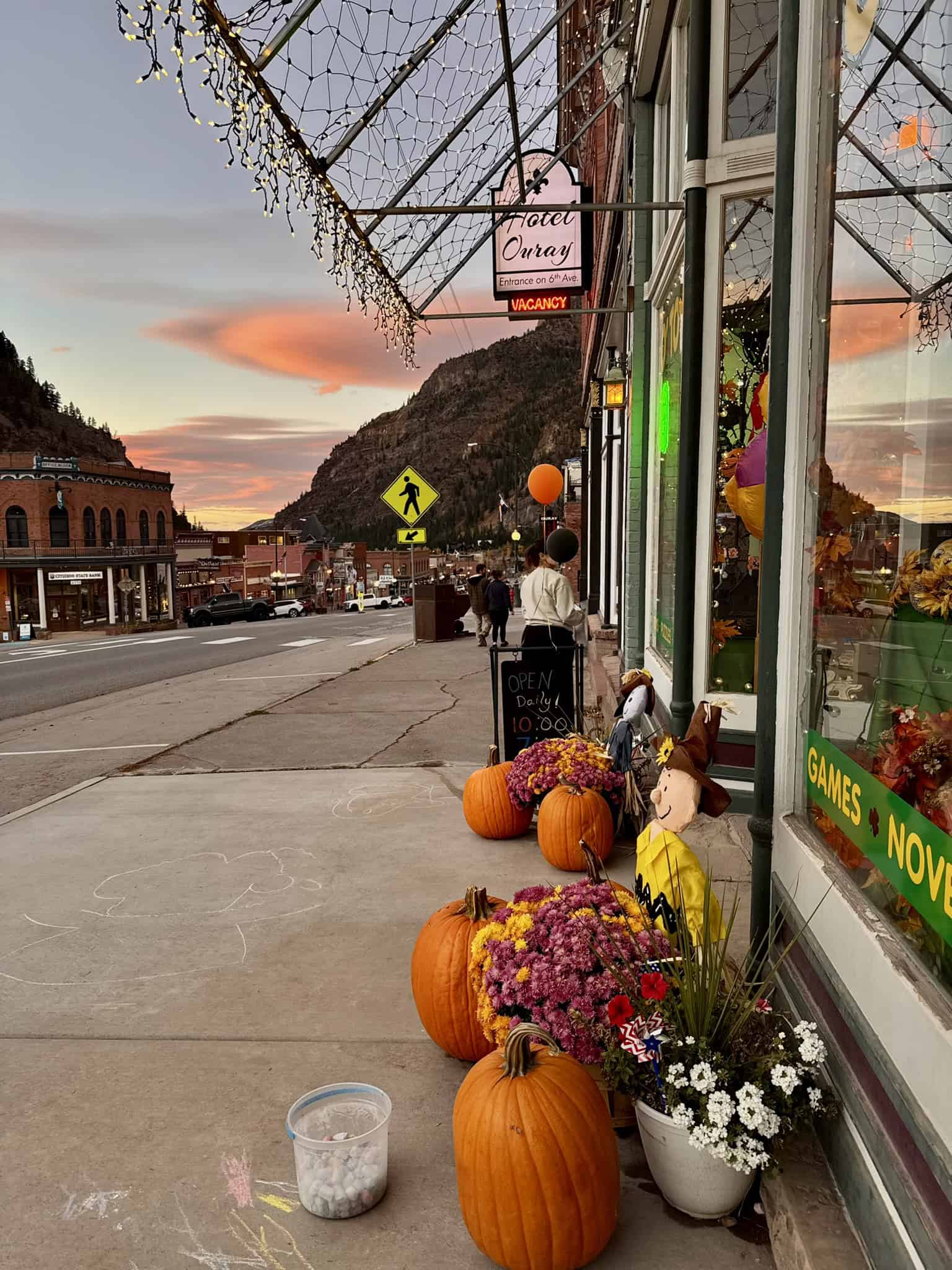 A charming walkway along Ouray Main Street, lined with pumpkins and flowers set against distant canyon walls and colorful clouds at sunset. 