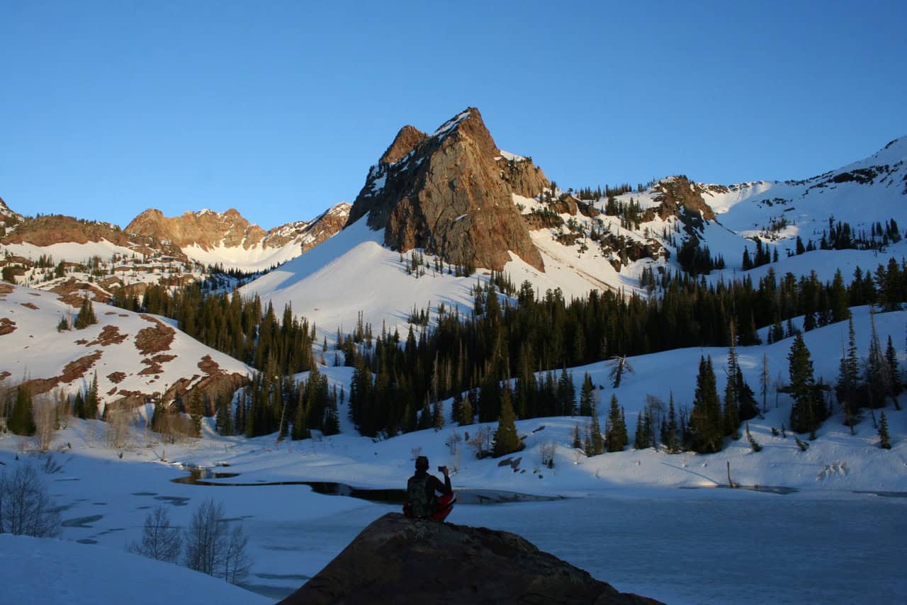 A hiker sits on a rock above a snowy Lake Blanche, with rugged mountain peaks and evergreen forests in the background