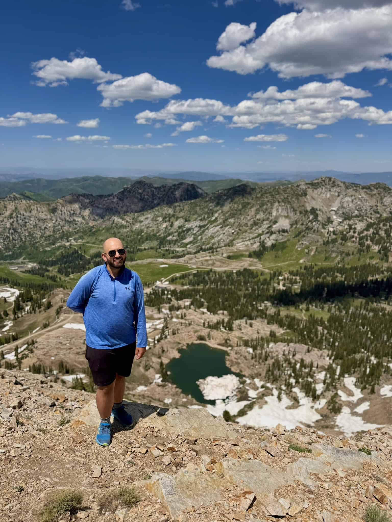 A man stands on a mountain summit with a wide view of a rugged landscape, including a small lake with chunks of ice, surrounded by green slopes and distant peaks under a blue sky.


