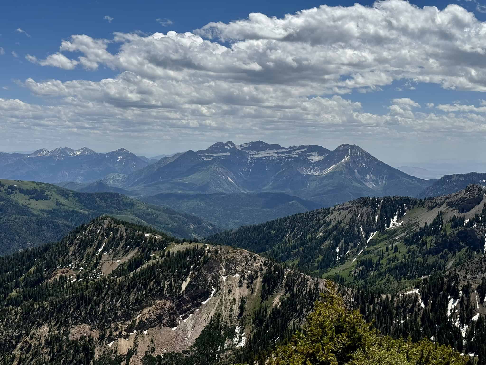 A scenic view from a Sugarloaf peak showing snow-dappled ridges, forested valleys, and the towering form of Mount Timpanogos in the distance beneath a partly cloudy sky.