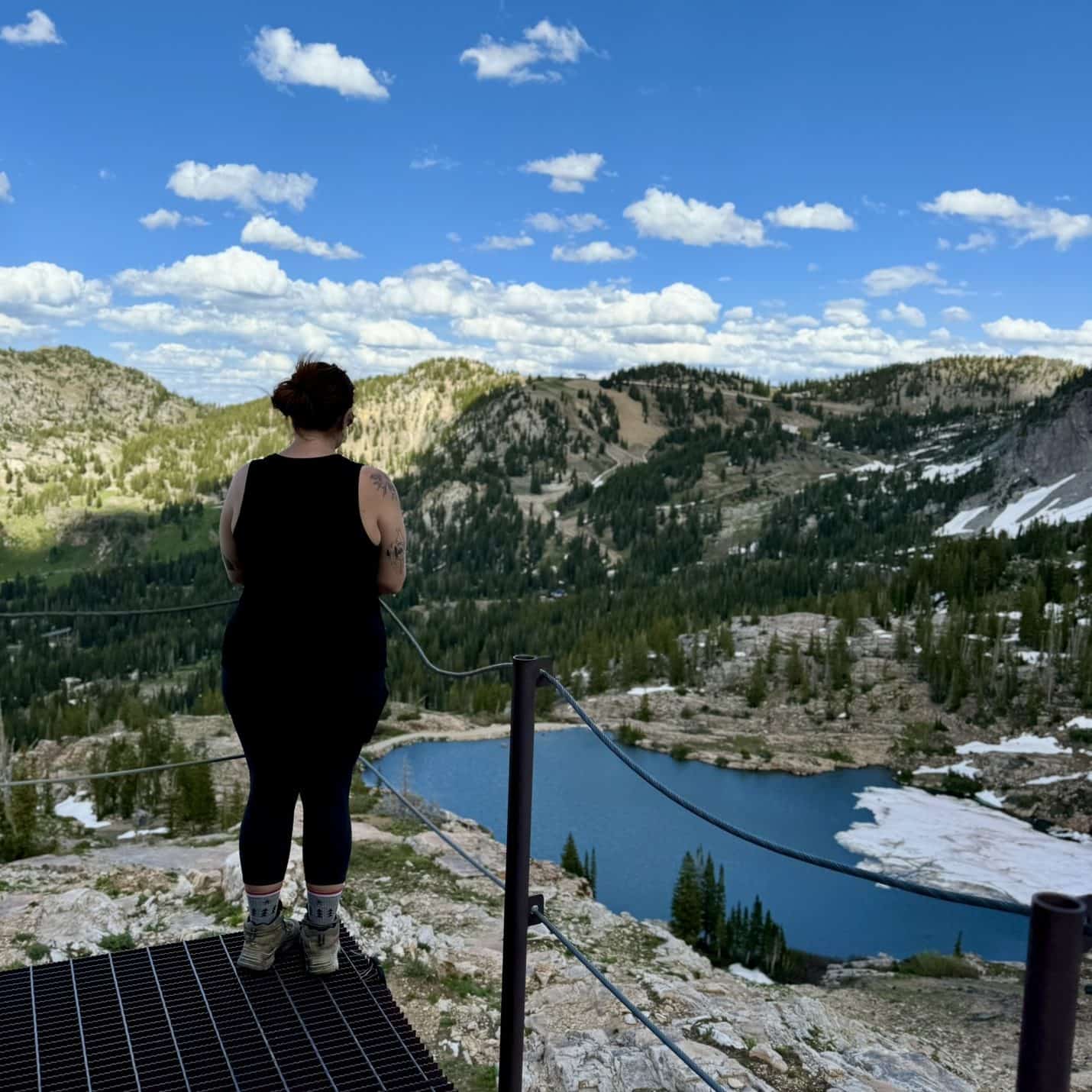 A woman stands on a metal platform overlooking Cecert Lake, a scenic hike in Little Cottonwood Canyon, with its deep blue waters nestled among forested mountains under a bright sky.