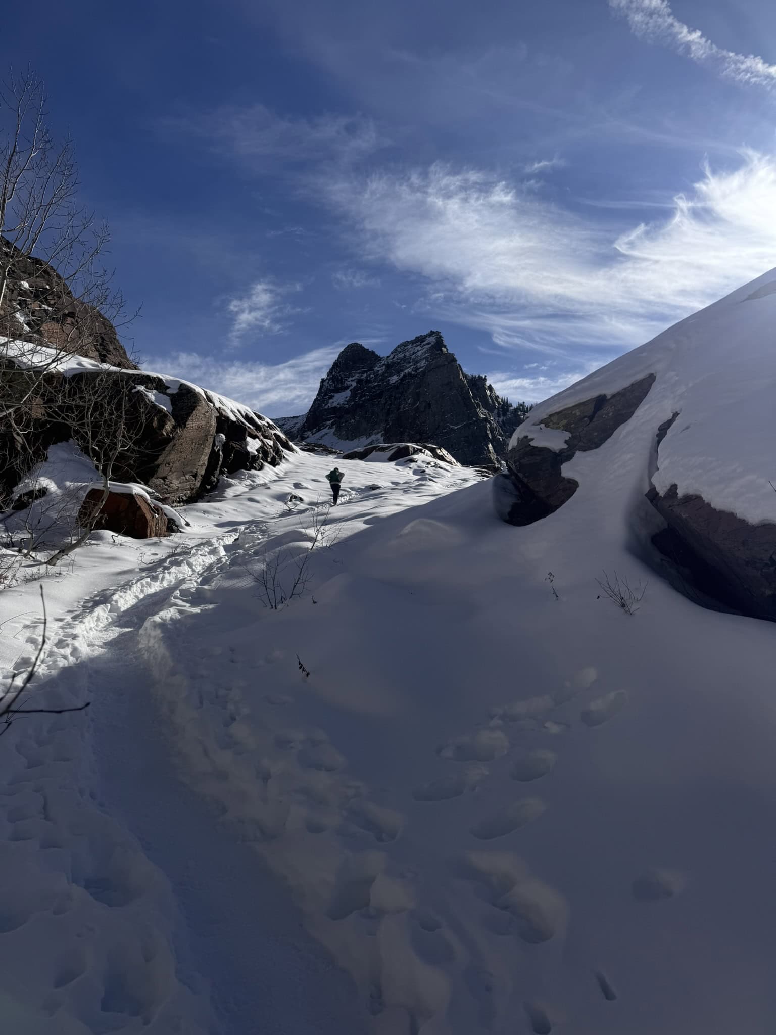 A hiker makes their way through deep snow along the Lake Blanche trail with Sundial Peak looming in the distance, under a bright blue sky
