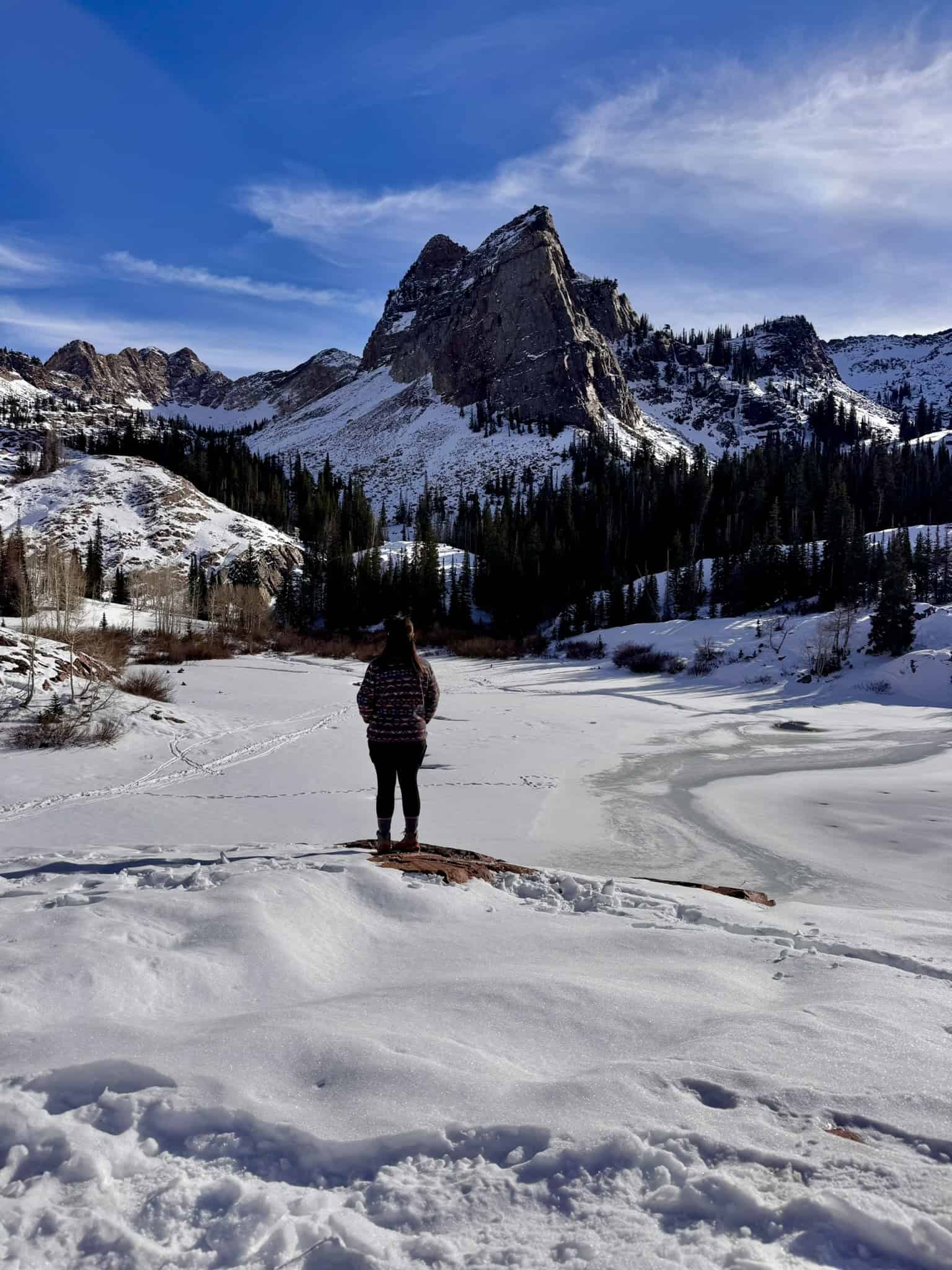 A serene view of Lake Blanche in winter, with a hiker standing above the frozen lake beneath a bright blue sky and towering snow-dusted mountains.



