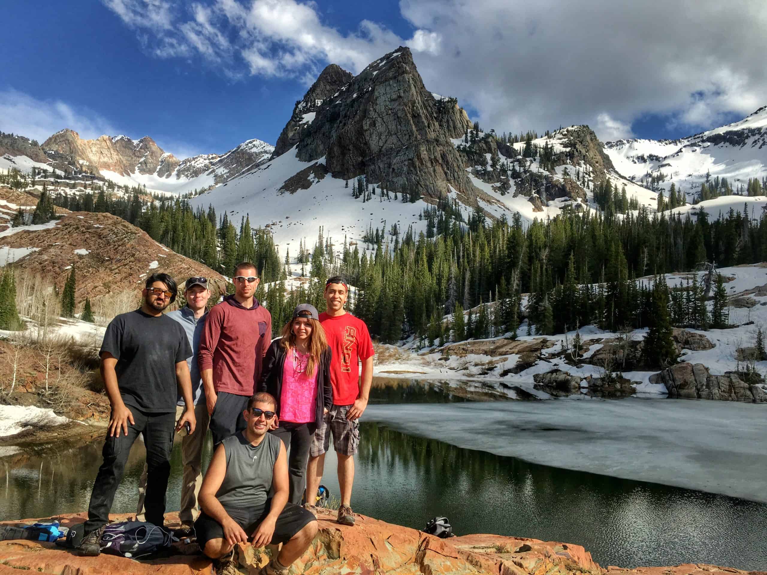 A group of hikers poses for a photo beside a partially frozen Lake Blanch, surrounded by snow-covered mountains and green trees  under a partially cloudy blue sky.

