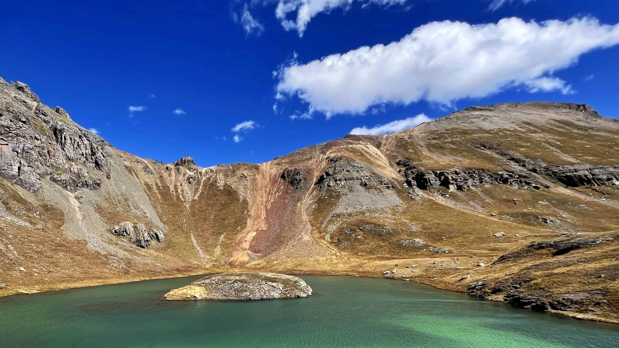 A vibrant emerald lake with a small island nestled in a mountain basin  with V2 peak looming to the right under partly cloudy skies