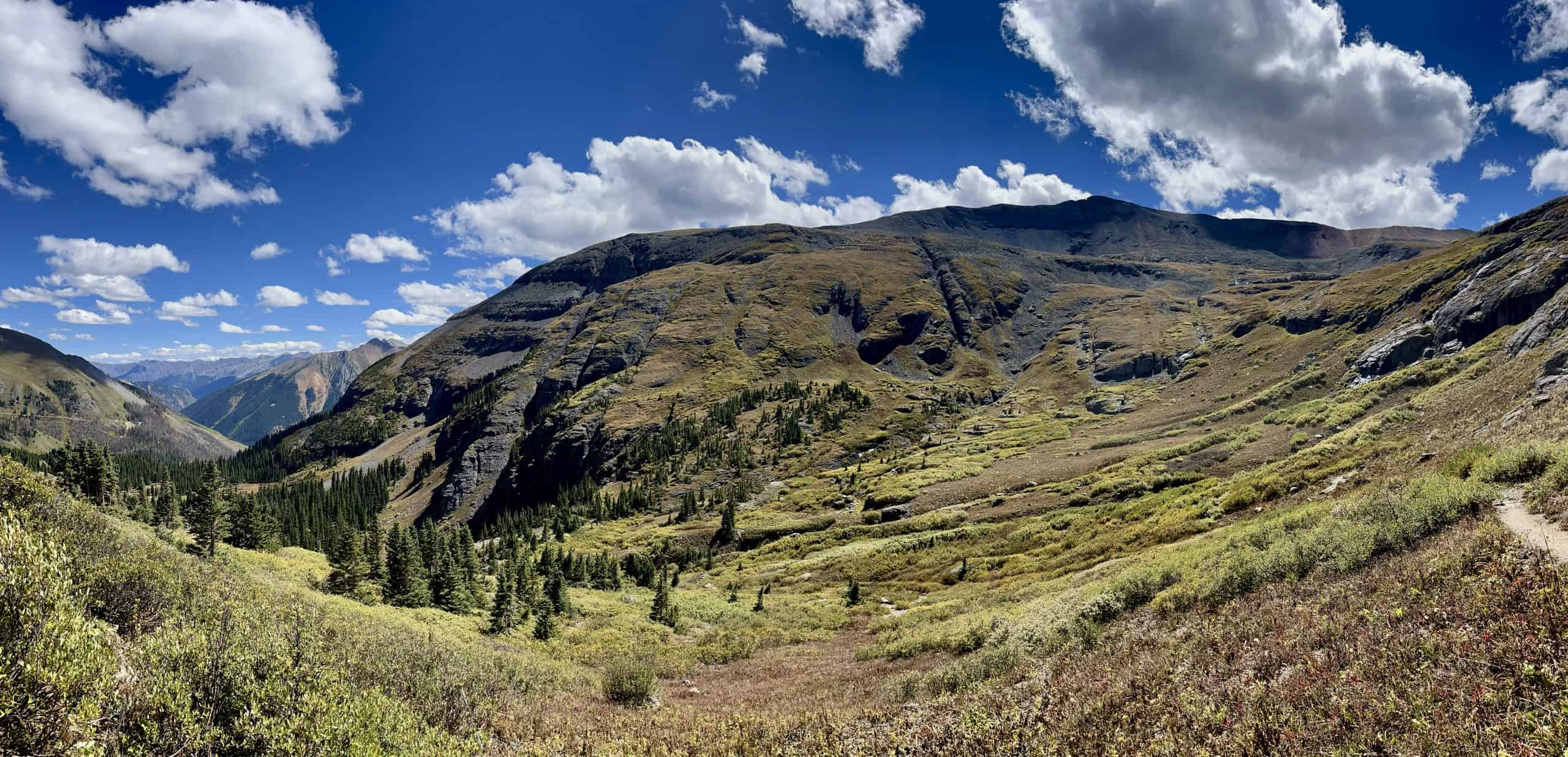 A panoramic view of high-altitude alpine tundra and rocky slopes leading toward distant mountain peaks under a bright blue sky with scattered clouds, on the descent from Ice Lake Colorado.

