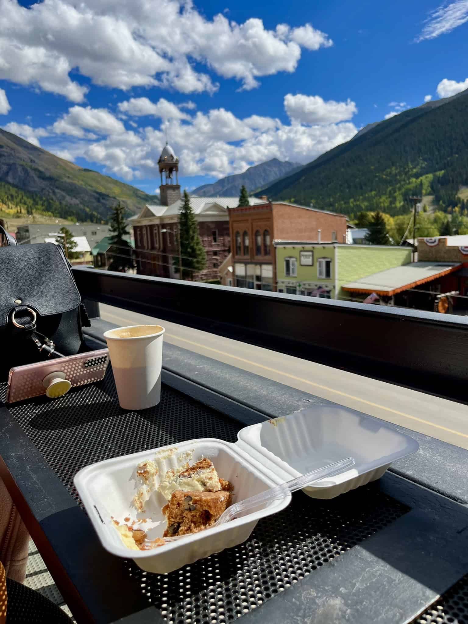 A coffee cup and carrot cake on a rooftop table with a scenic view of Silverton’s buildings and mountains under a partly cloudy blue sky, 