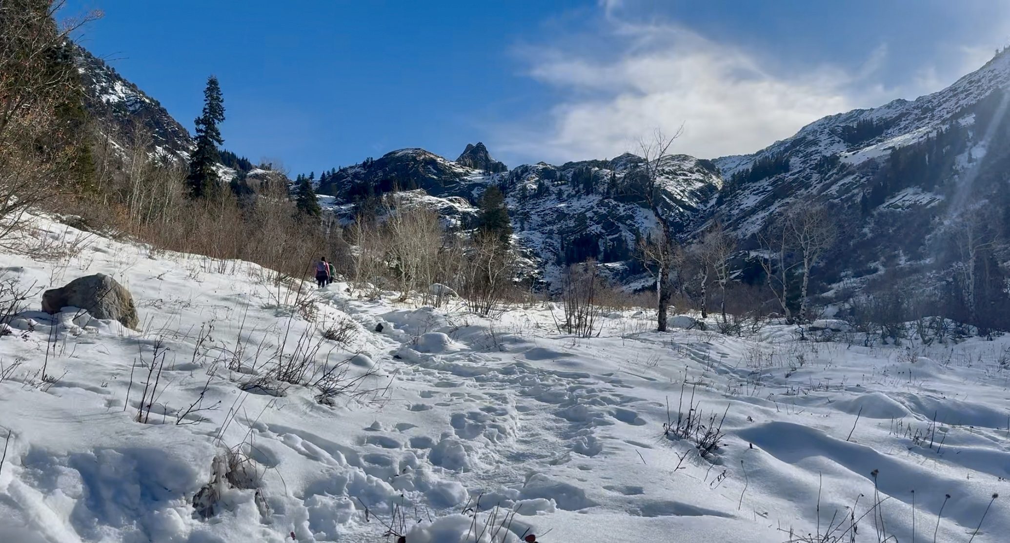 A snowy trail winds through a winter meadow along the Lake Blanche Trail, with hikers visible in the distance under a bright blue sky.