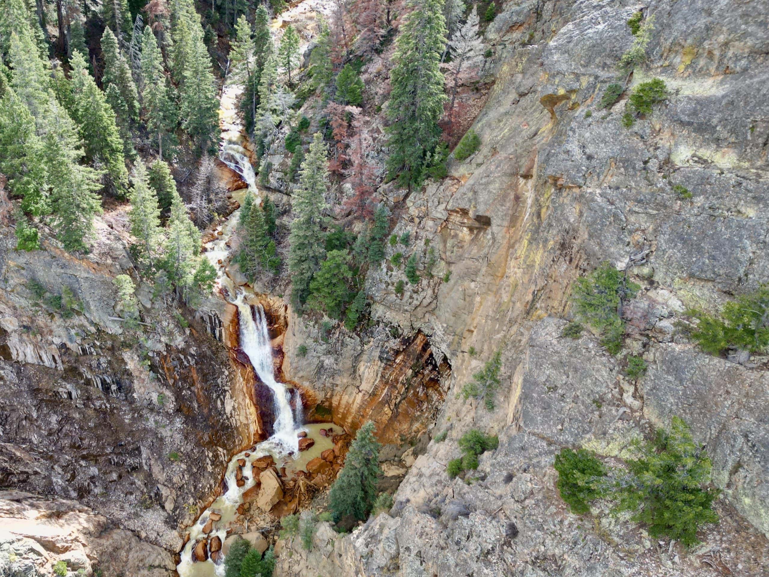 A aerial view of Waterfall Wall, a dramatic rock face with a cascading waterfall surrounded by evergreen trees and rugged cliffs, showcasing an exciting climbing area and one of the many best things to do in Ouray
