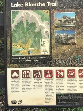 A trailhead sign for the Lake Blanche Trail, featuring a detailed map of the route, elevation gain of 2,720 feet (898 m), and distance of 2.8 miles (4.5 km) to the lake, along with scenic images of the surrounding mountain landscape.