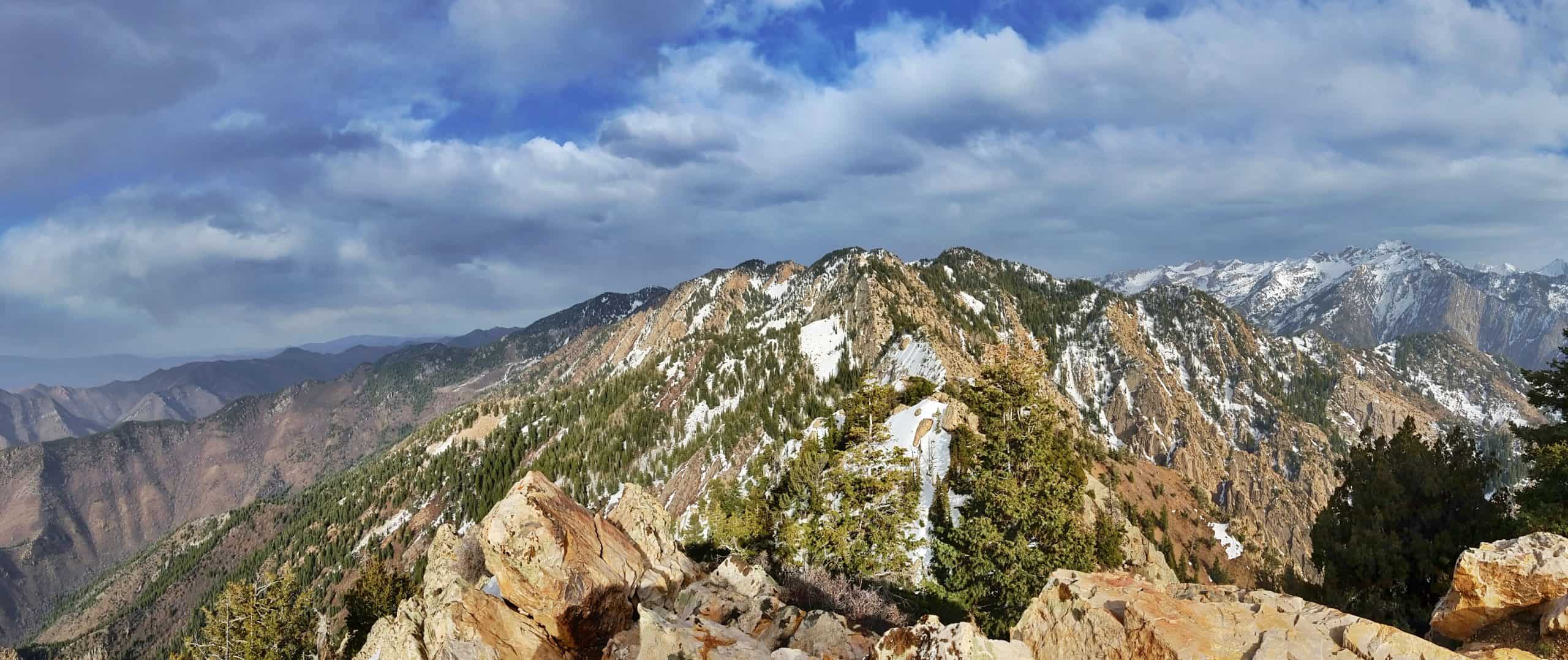 From the summit of Mount Olympus, rocky outcrops in the foreground frame sweeping mountain ridges dotted with evergreens and snow under a partly cloudy sky,
