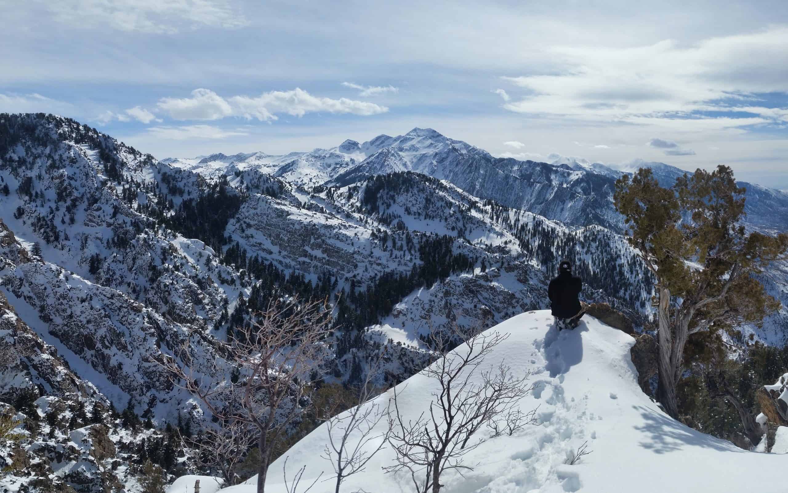 A hiker sits on a snow-covered rock with a snowy view of the Wasatch Mountains under a partly cloudy sky from the saddle of Mount Olympus. 

