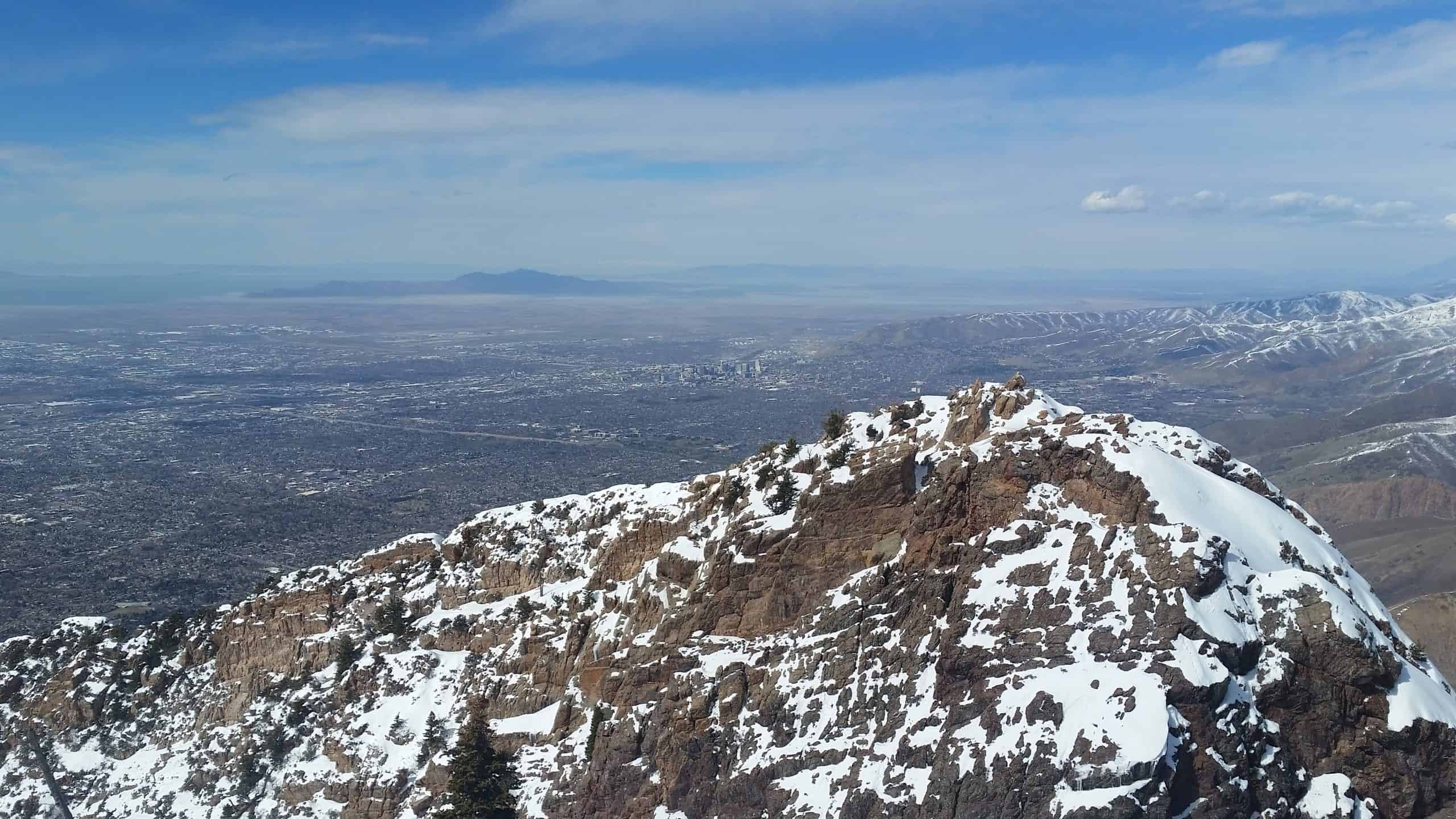A snow-dusted ridge dominates the foreground while overlooking the sprawling Salt Lake Valley and distant peaks under a clear blue sky, from the summit of Mount Olympus, Utah. 