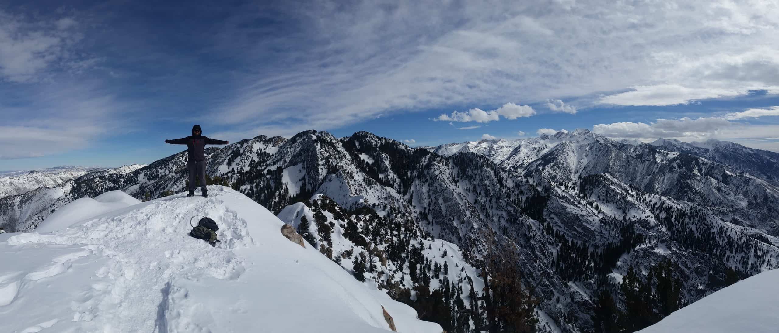 A hiker stands with arms outstretched on the snow-covered Mount Olympus summit, with vast, rugged snowcapped mountain ranges in the background, under a bright sky streaked with clouds, 