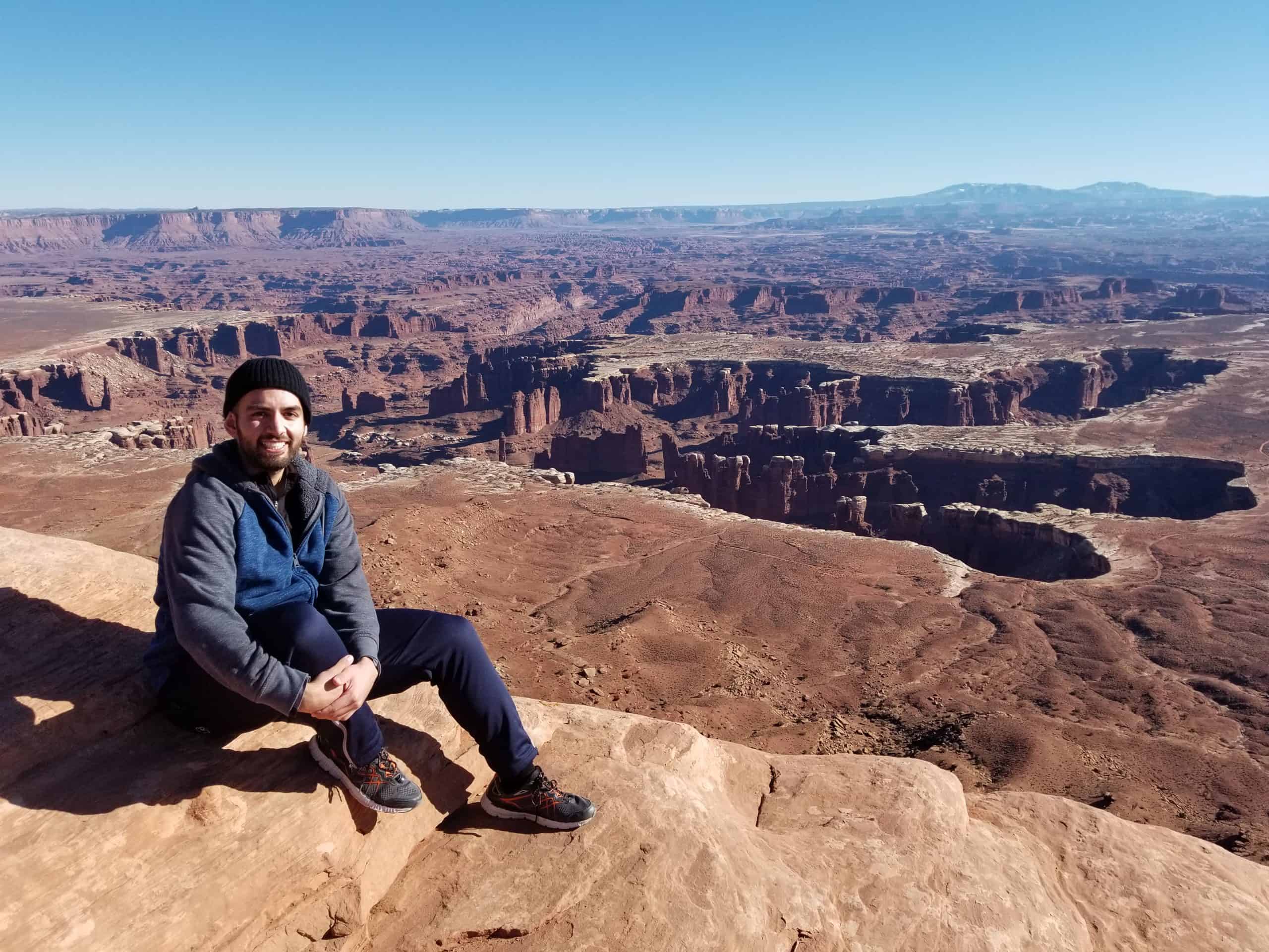 A man sitting and smiling towards the camera, with a stunning vast view of Canyonlands National Park in the background under a blue sky. 
