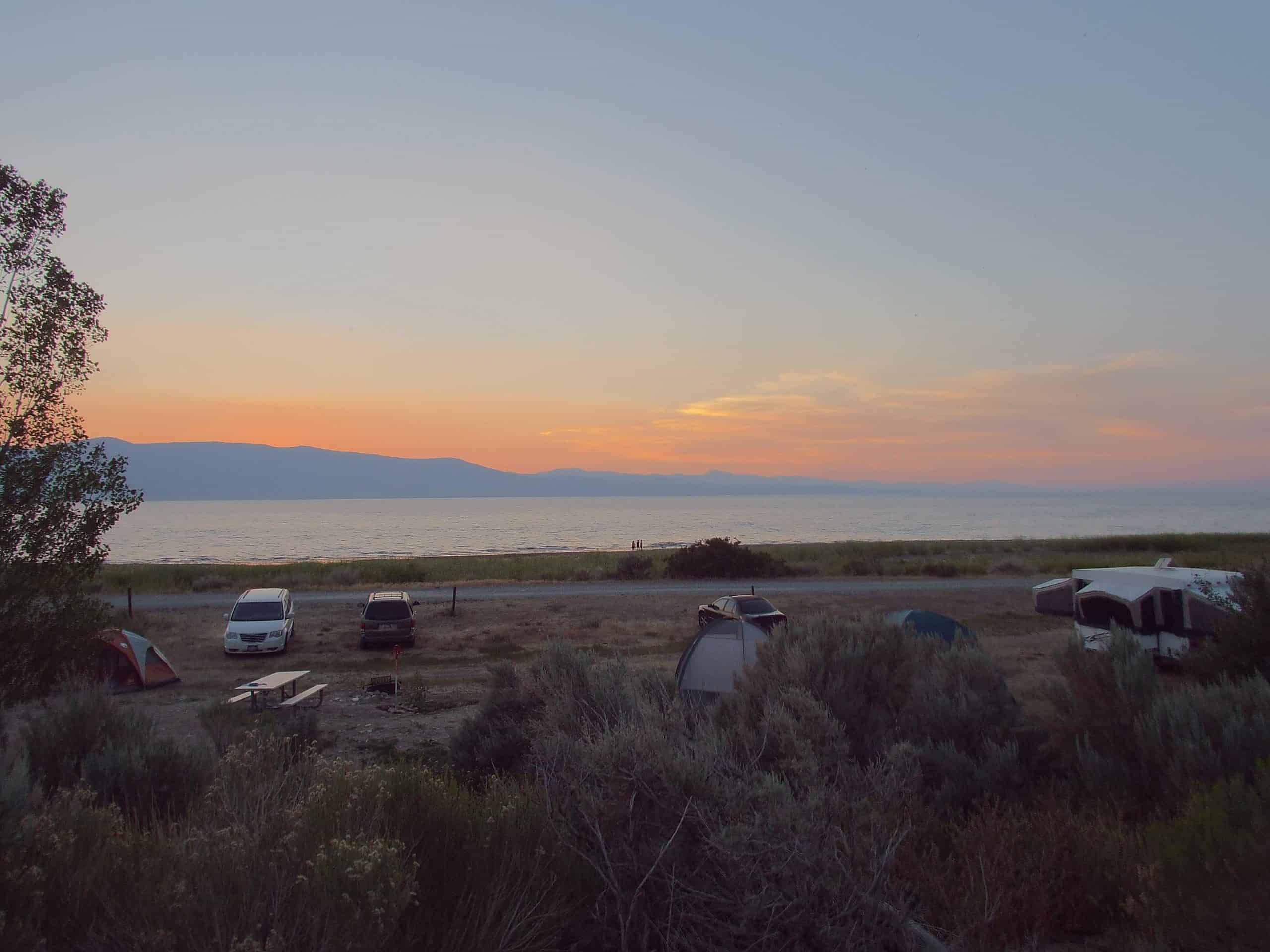 Cisco Beach Campground at sunset, with campers and vehicles set up near the serene shoes of Bear Lake, and distant mountain peaks