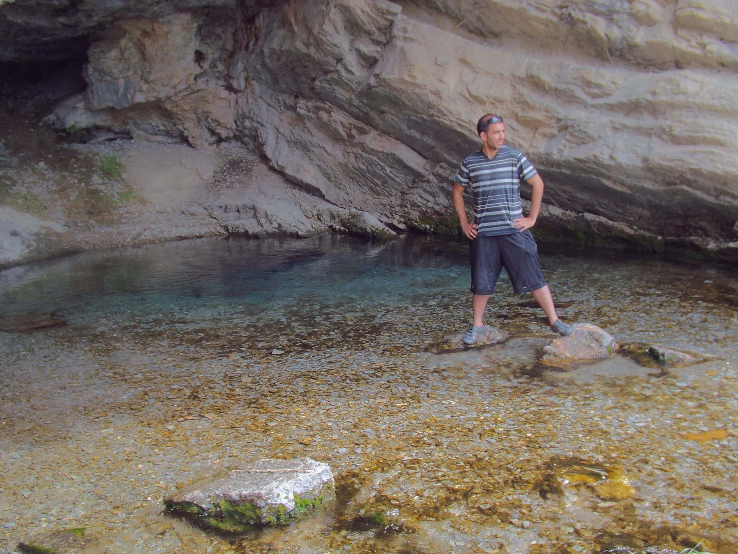 A man standing on two rocks surrounded by clear water at Rick's Spring in Logan Canyon, set against  rocky cliffs