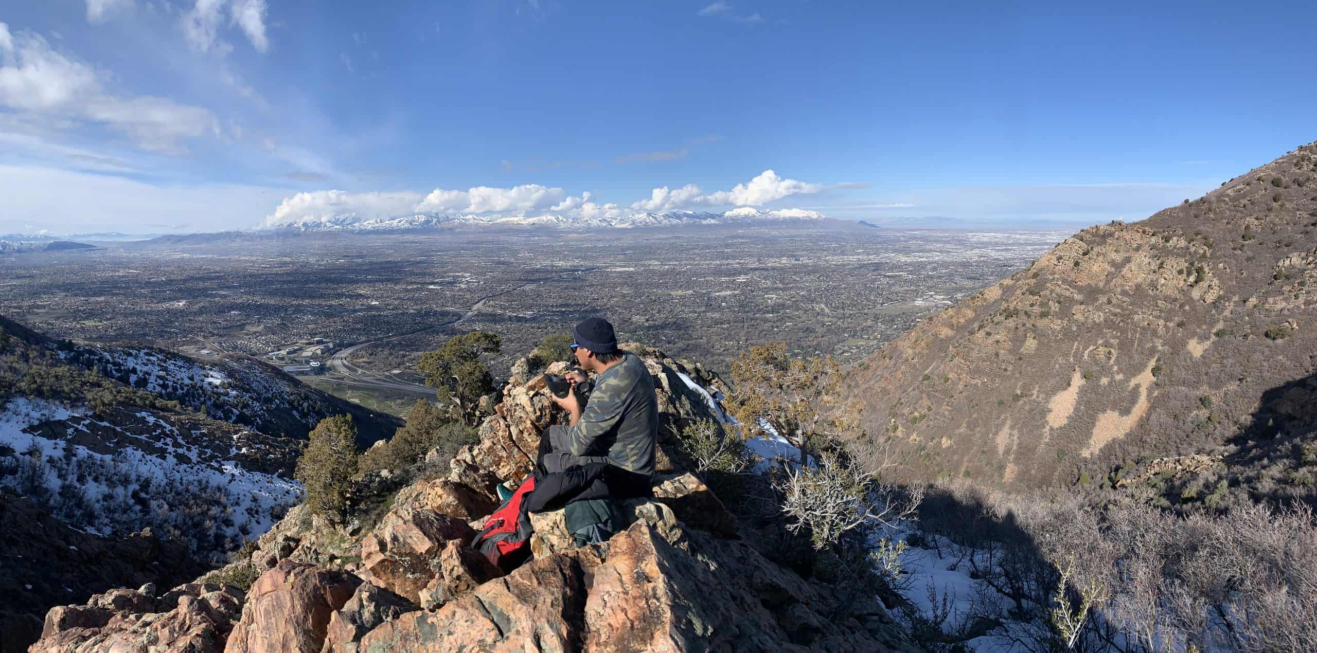 A hiker rests on a rocky outcrop gazing across the Salt Lake Valley toward distant snowcapped mountain peaks, a scenic midway break on the Mount Olympus Hike in Utah