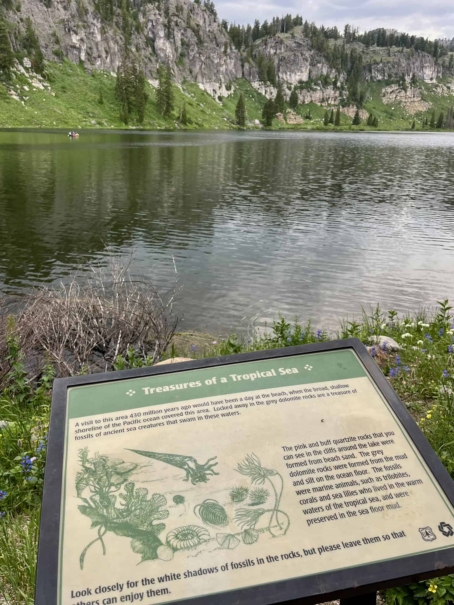 An interpretive sign about ancient sea life stands before a calm Tony Grove Lake with rocky cliffs in the backdrop. 