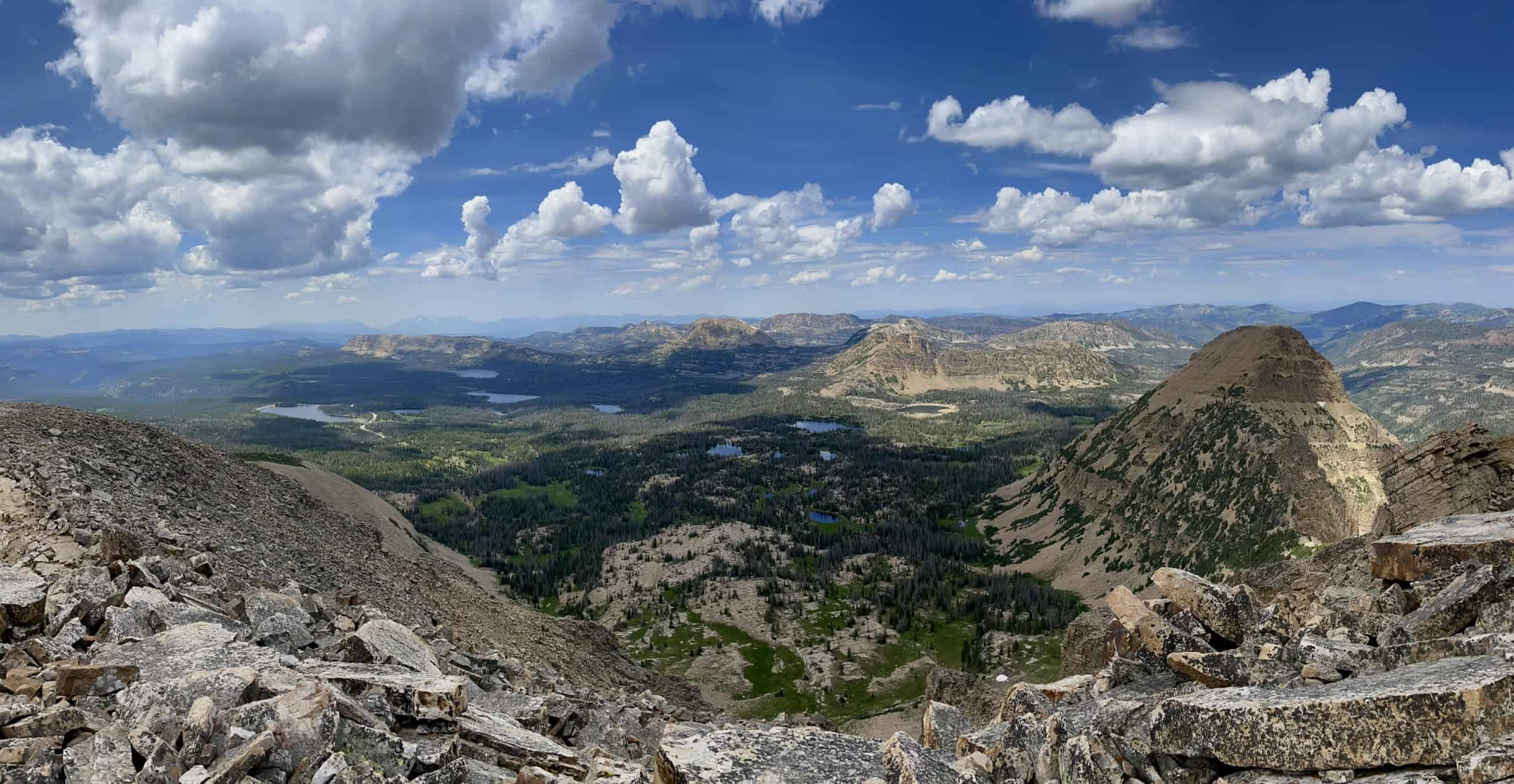 A panoramic view from the summit of the Bald Mountain hike in Utah, showcasing numerous mountains and lakes under scattered clouds and blue skies.

