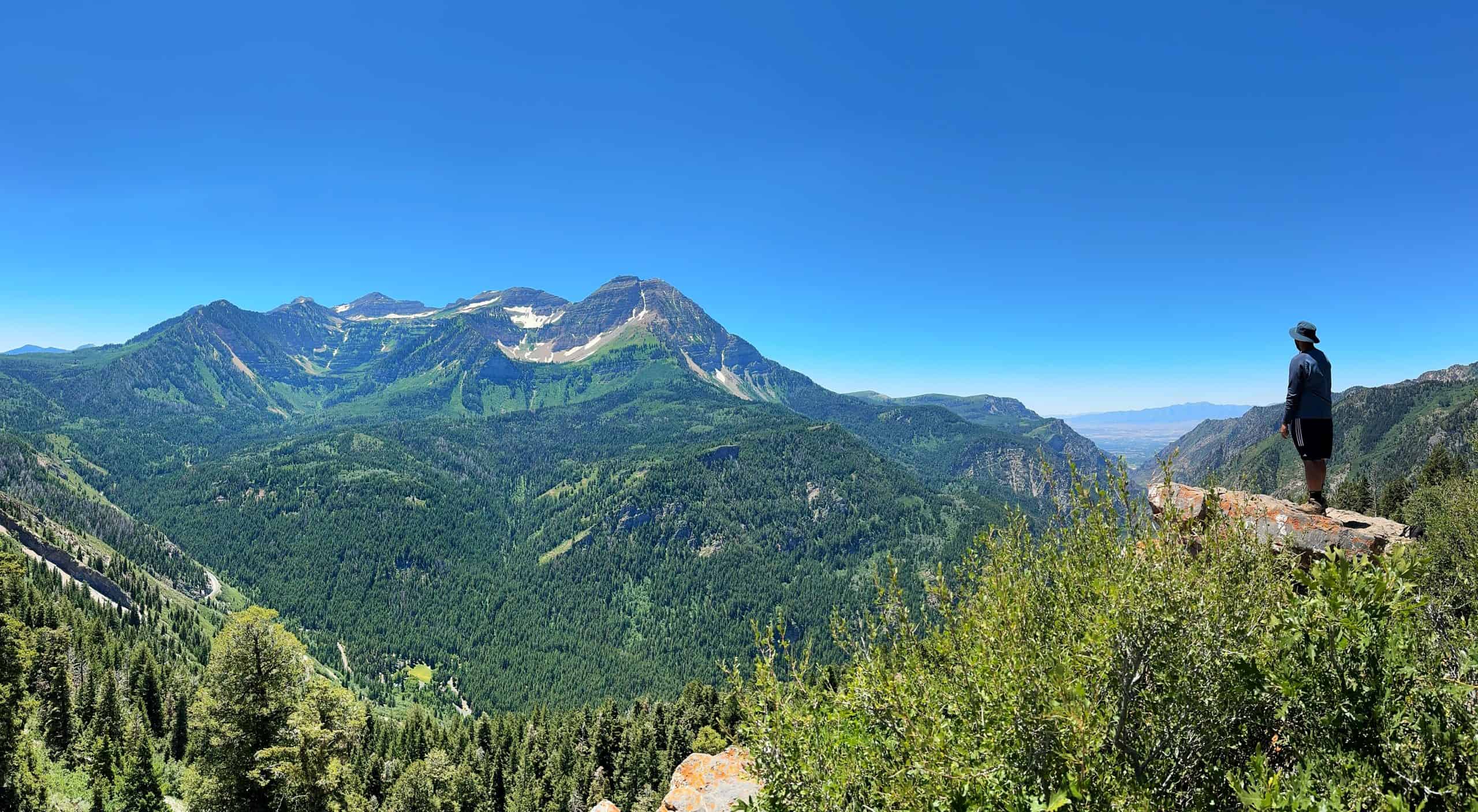 A hiker stands atop a rocky outcrop gazing towards the majestic Mount Timpanogos off in the distance from Pine Hollow Peak, under a bright blue sky

