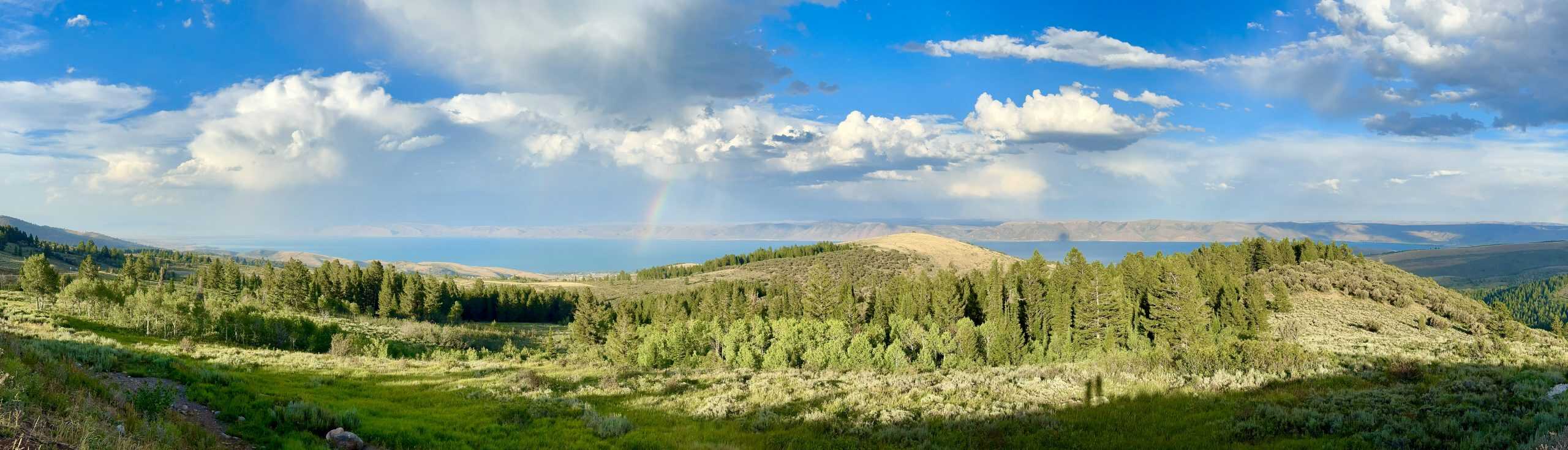 A scenic view of Bear Lake from a grassy hillside under a dramatic sky with clouds and a faint rainbow