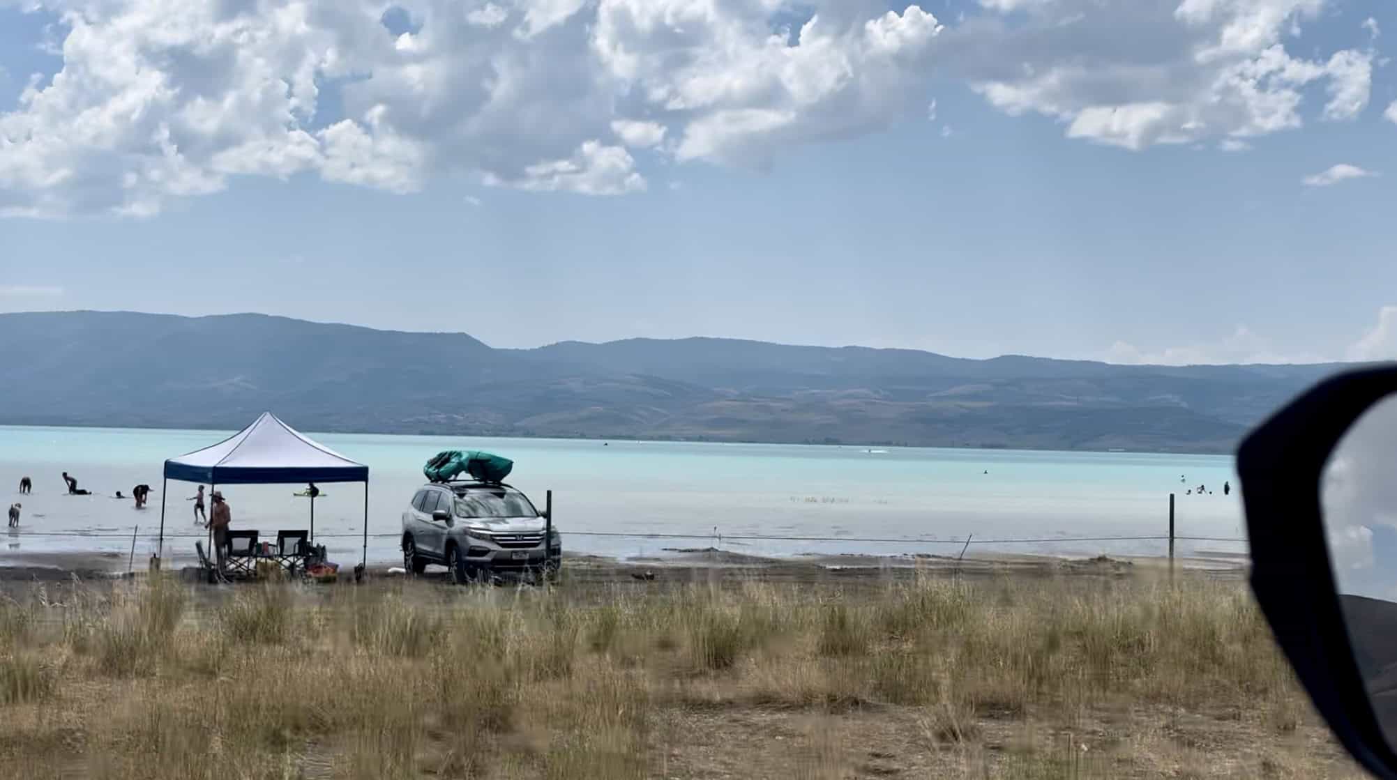 An open view of the Turquoise waters of the North Shore of Bear Lake, with distant mountain peaks, and cars parked in the foreground. 