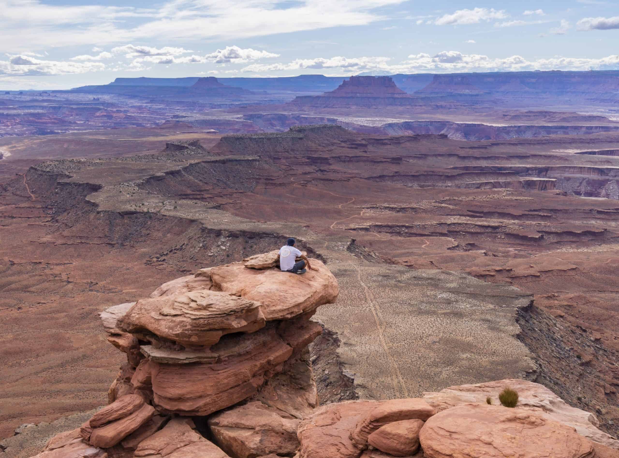  A hiker rests on a flat-topped sandstone formation overlooking vast canyon floors and distant mesas under a partly cloudy sky, while hiking in Canyonlands National Park.
