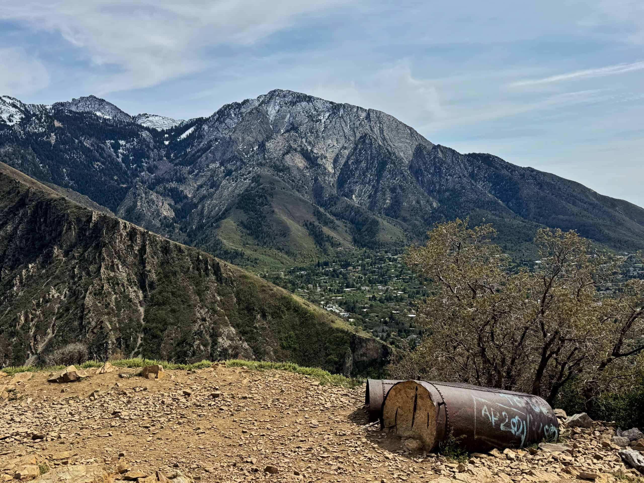 A weathered pipeline on a dirt travel sit against the steep, rugged and snow-dusted north face of Mount Olympus rising above a green valley. 