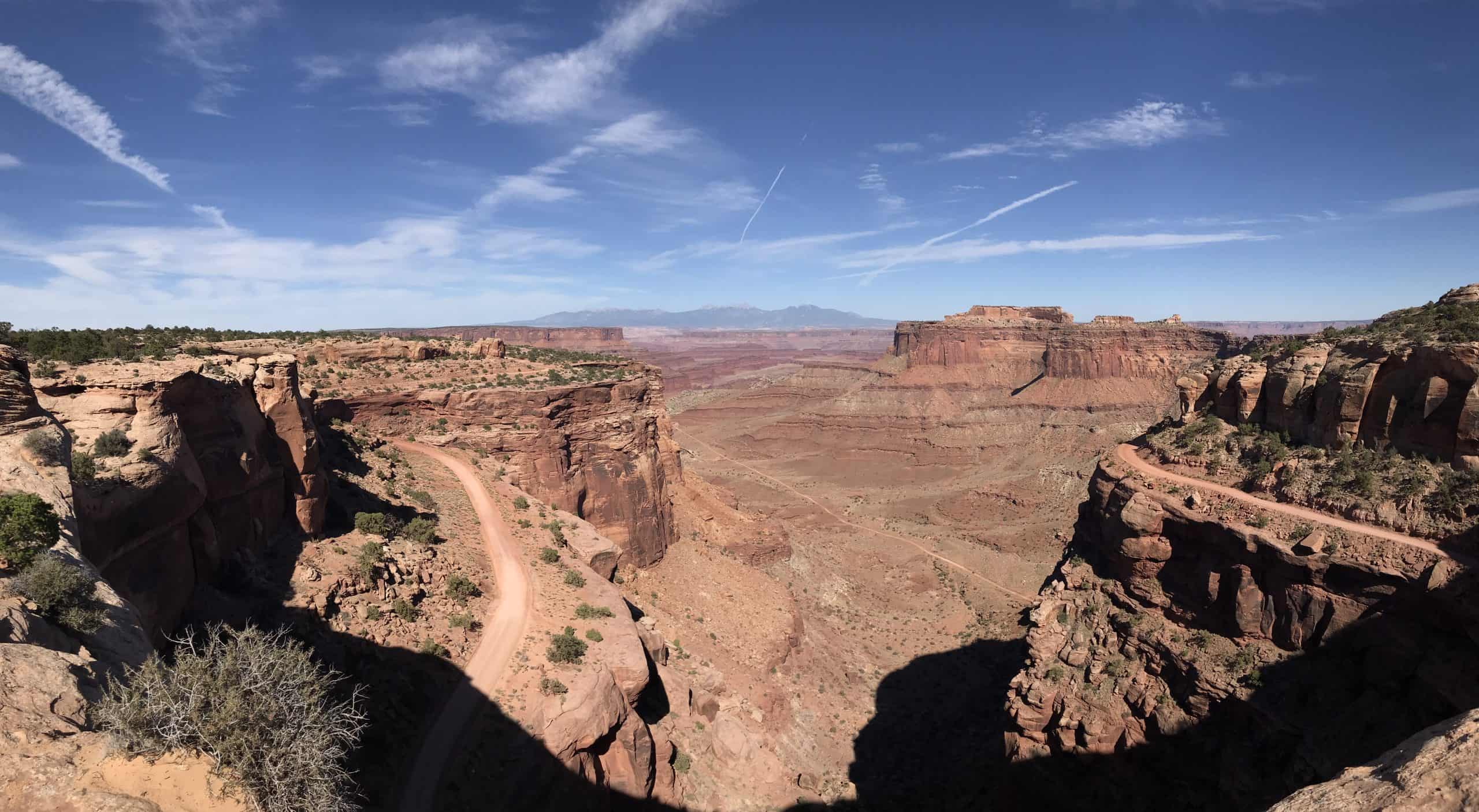 A winding dirt road traces the rim of a vast red rock canyon under a bright blue sky from the Shafer Canyon Overlook in Canyonlands National Park.