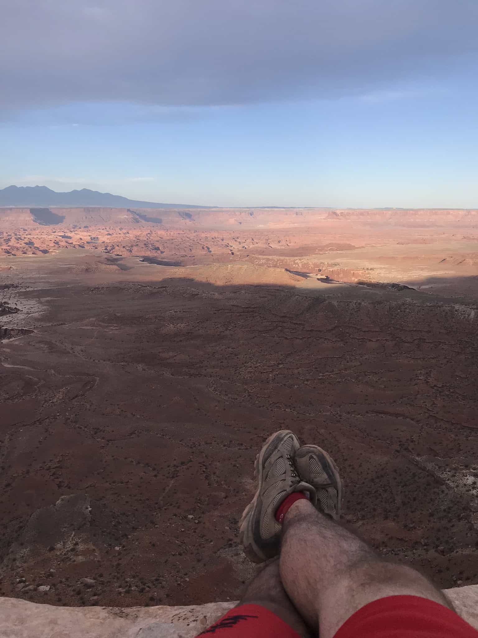 A man lays, resting his boots on this cliff edge, revealing a sweeping view of Gooseberry Canyon bathed in sunlight while hiking in Canyonlands National Park.