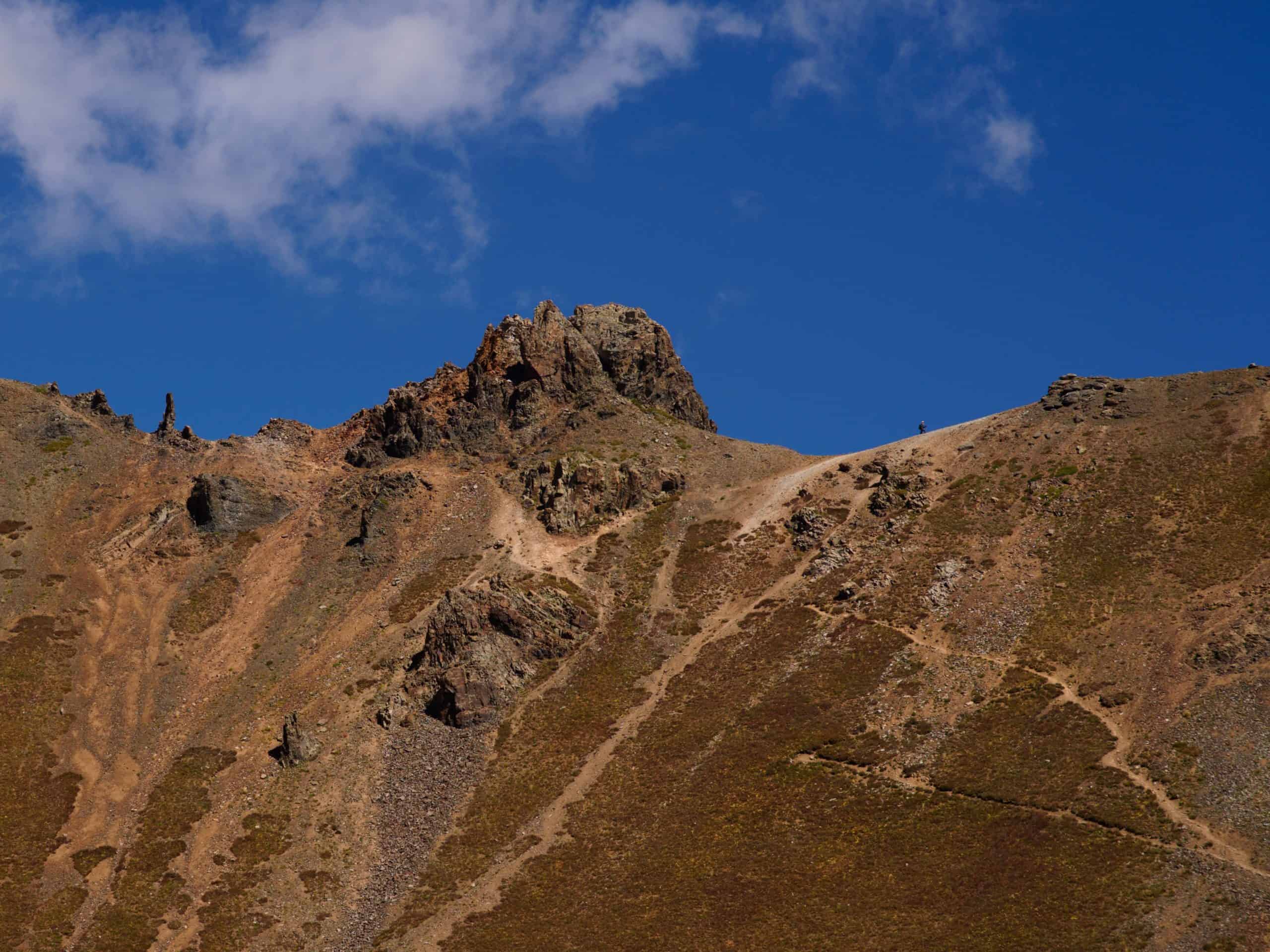A solitary hiker traverses a jagged, rocky ridge under a bright blue sky with wispy clouds, enroute to V2 Peak near Ice Lake, Colorado.


