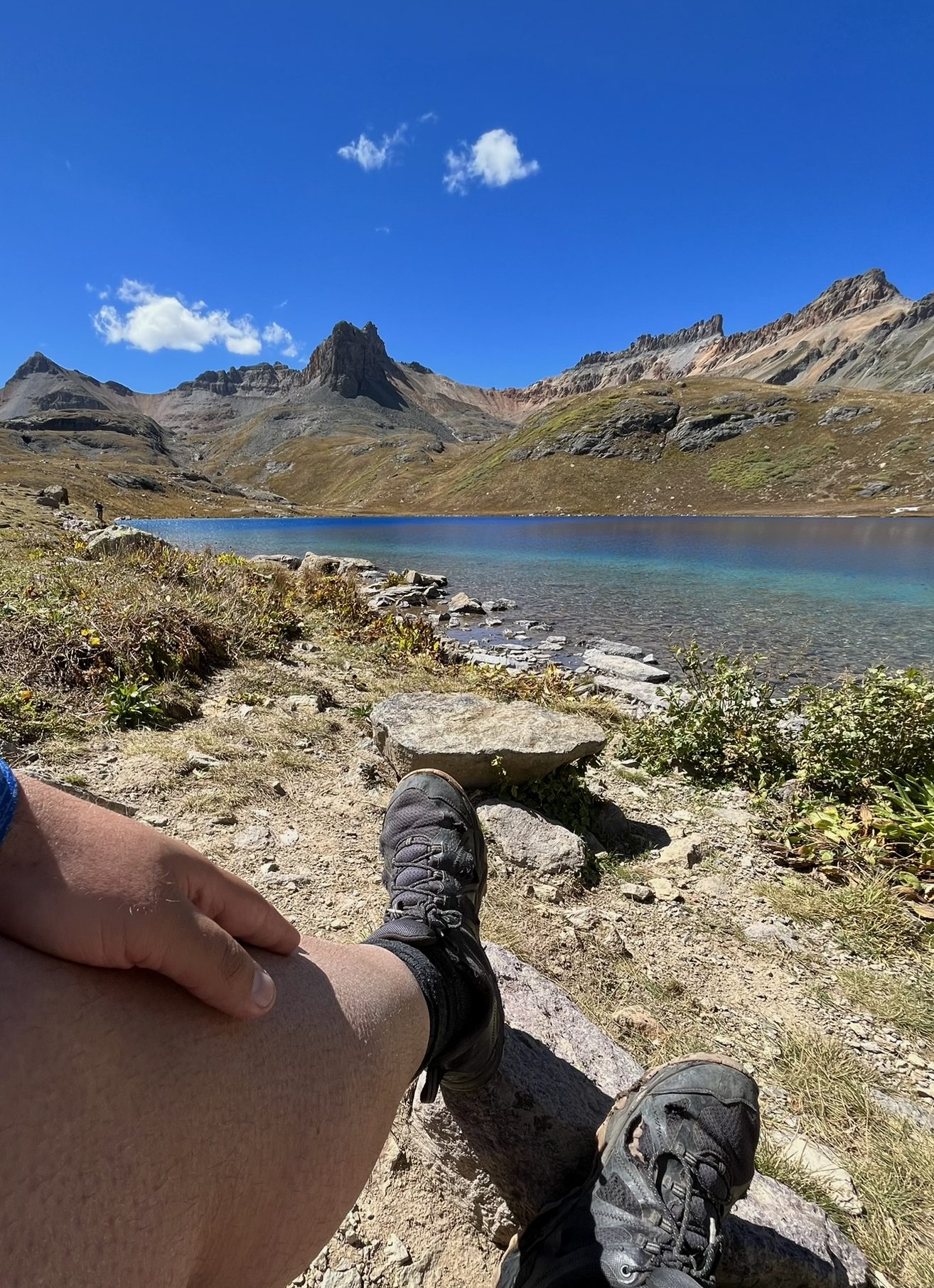 A close-up view of a hiker's black hiking boot resting on rocky ground, looking out across the clear blue waters of Ice Lake Colorado.

