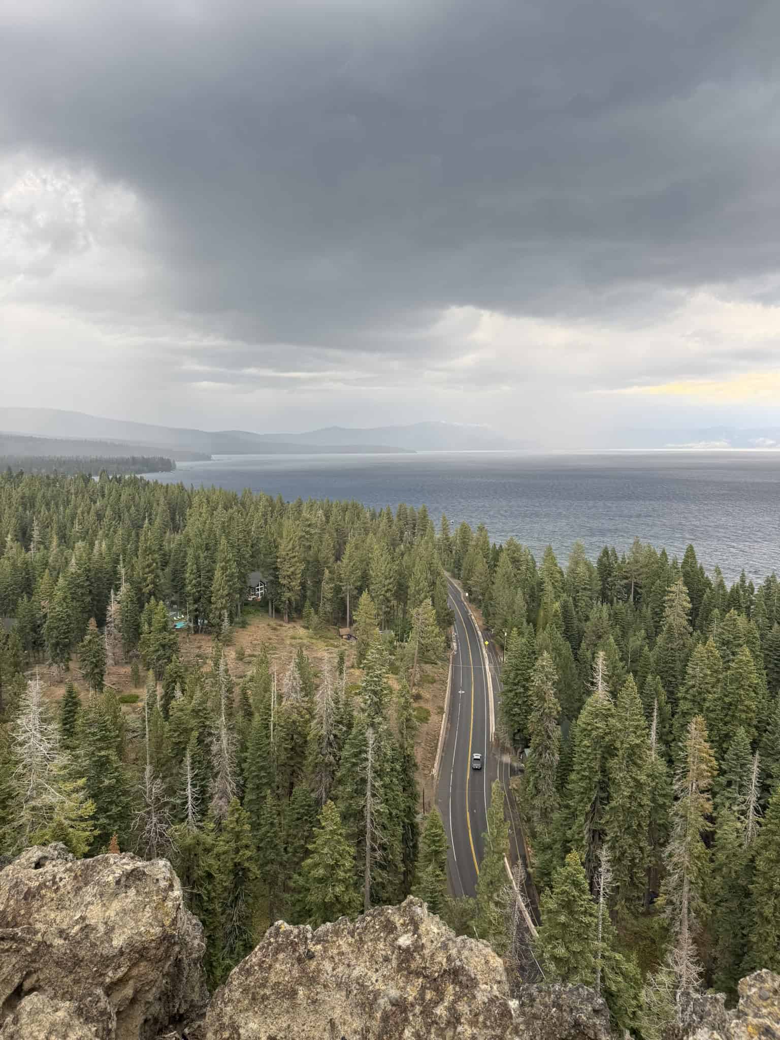 A winding road cuts through a dense pine forest toward Lake Tahoe, viewed from Eagle Rock under dramatic storm clouds.