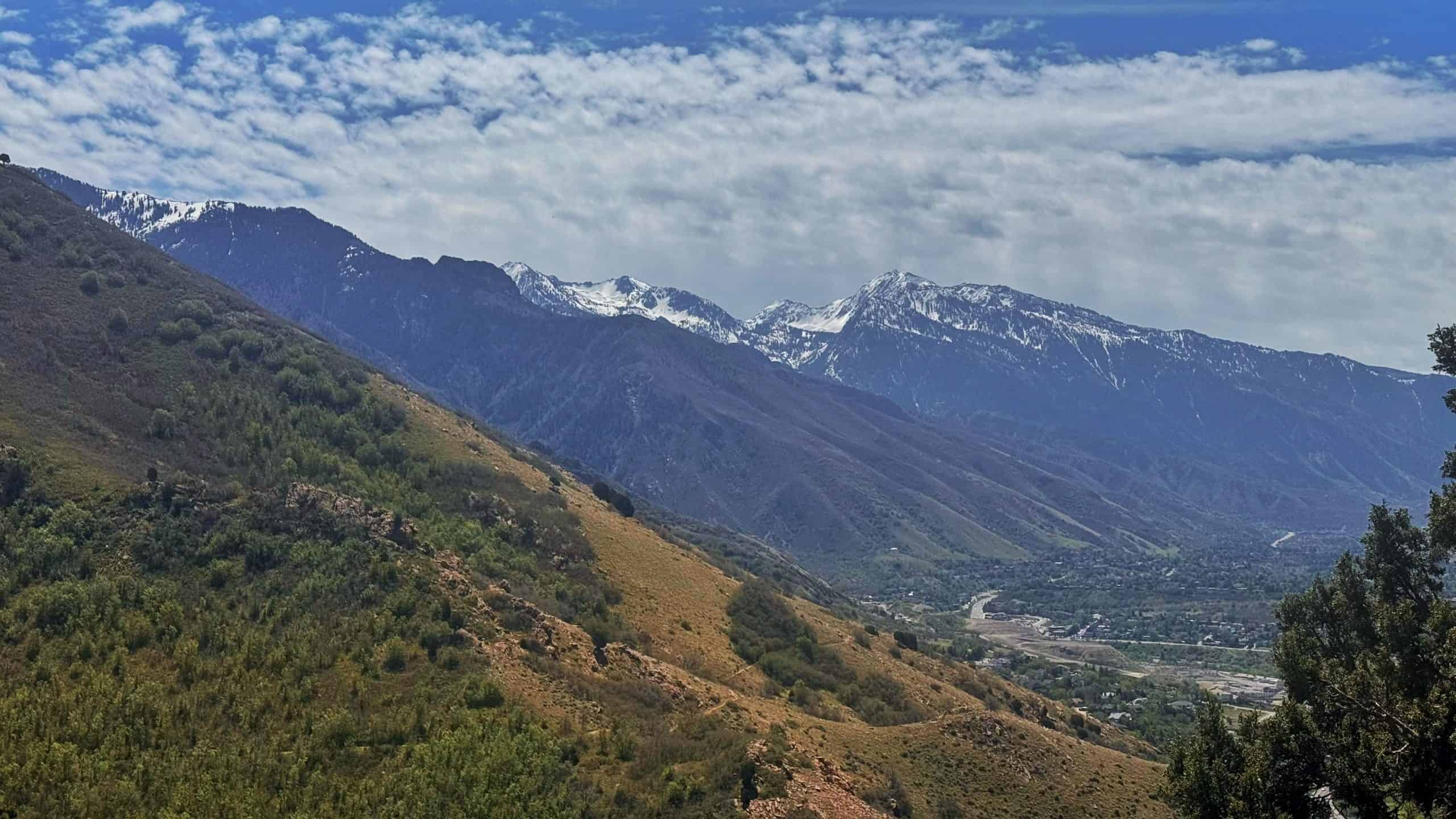 Snow-capped Lone Peak and Thunder Mountain rise above rolling green slopes under a partly cloudy sky,