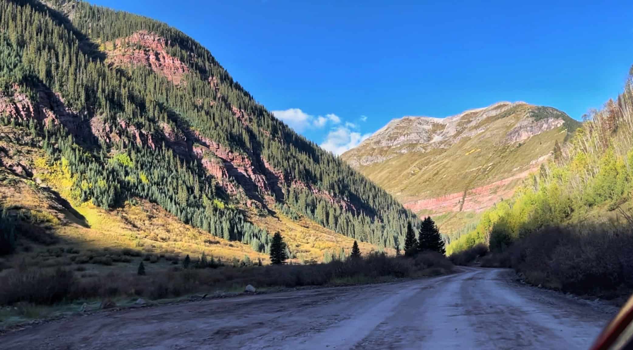 A scenic dirt road winds through a valley lined with evergreen trees and striking red rock cliffs under a blue sky, capturing the drive to the Ice Lake Colorado trailhead . 

