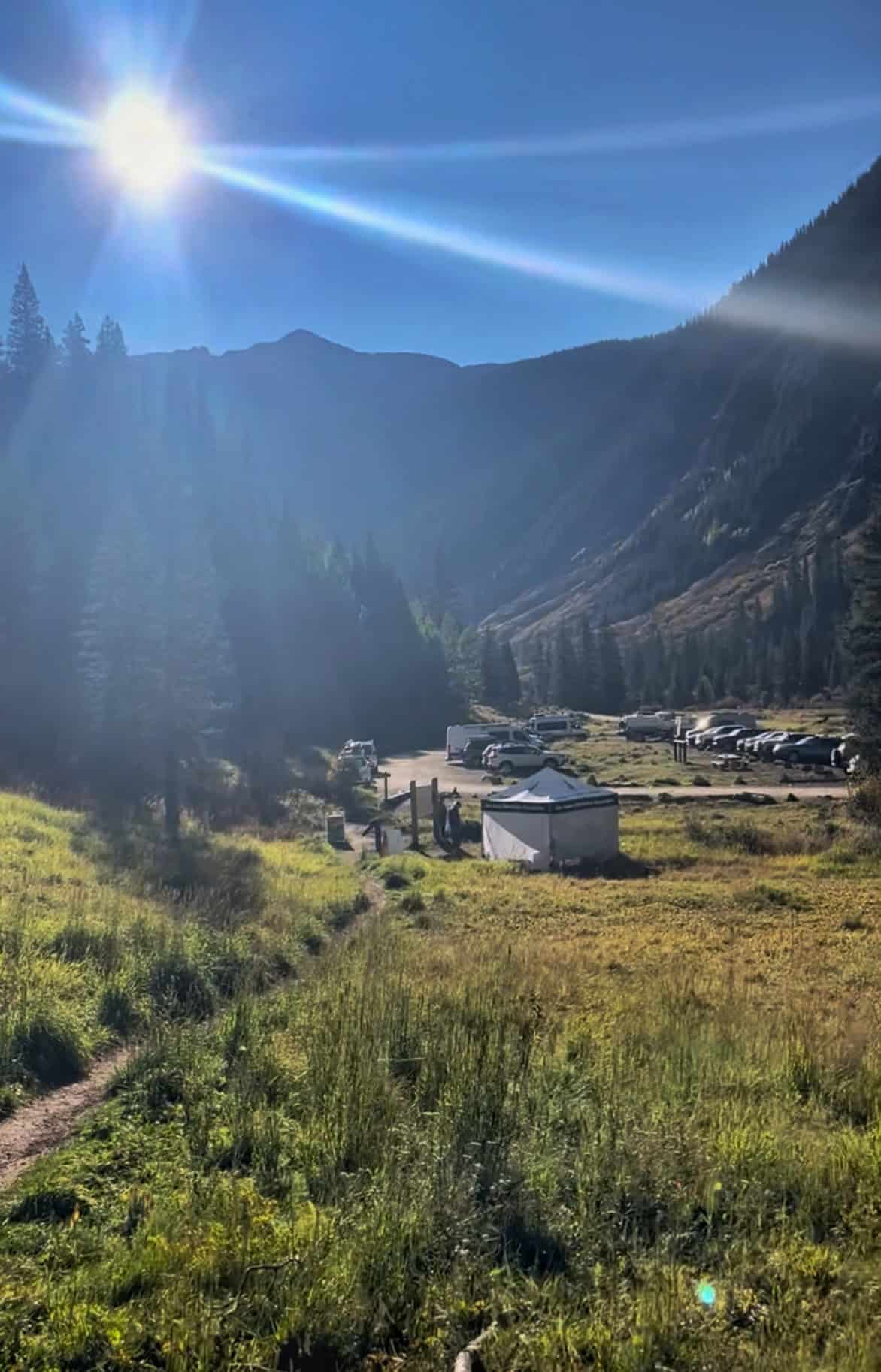 A sunny view of a gravel parking lot with a row of vehicles and a small white volunteer tent set against a backdrop of dark pine forests and mountain shadows at the trailhead for Ice Lake, Colorado.

