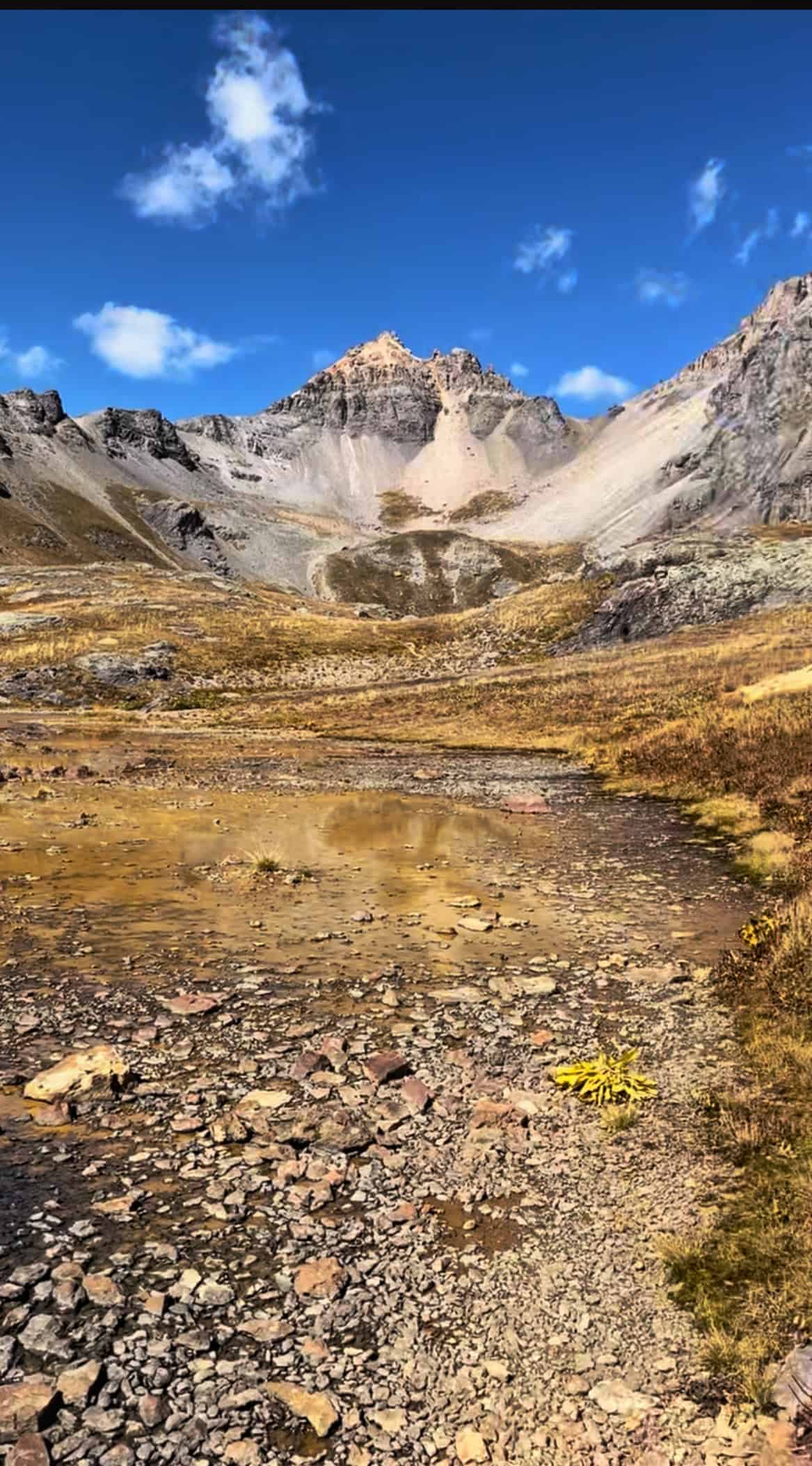 A small pond surrounded by tan grassy meadows leading to distant rugged peaks under a bright blue sky, enroute to Island Lake, Colorado