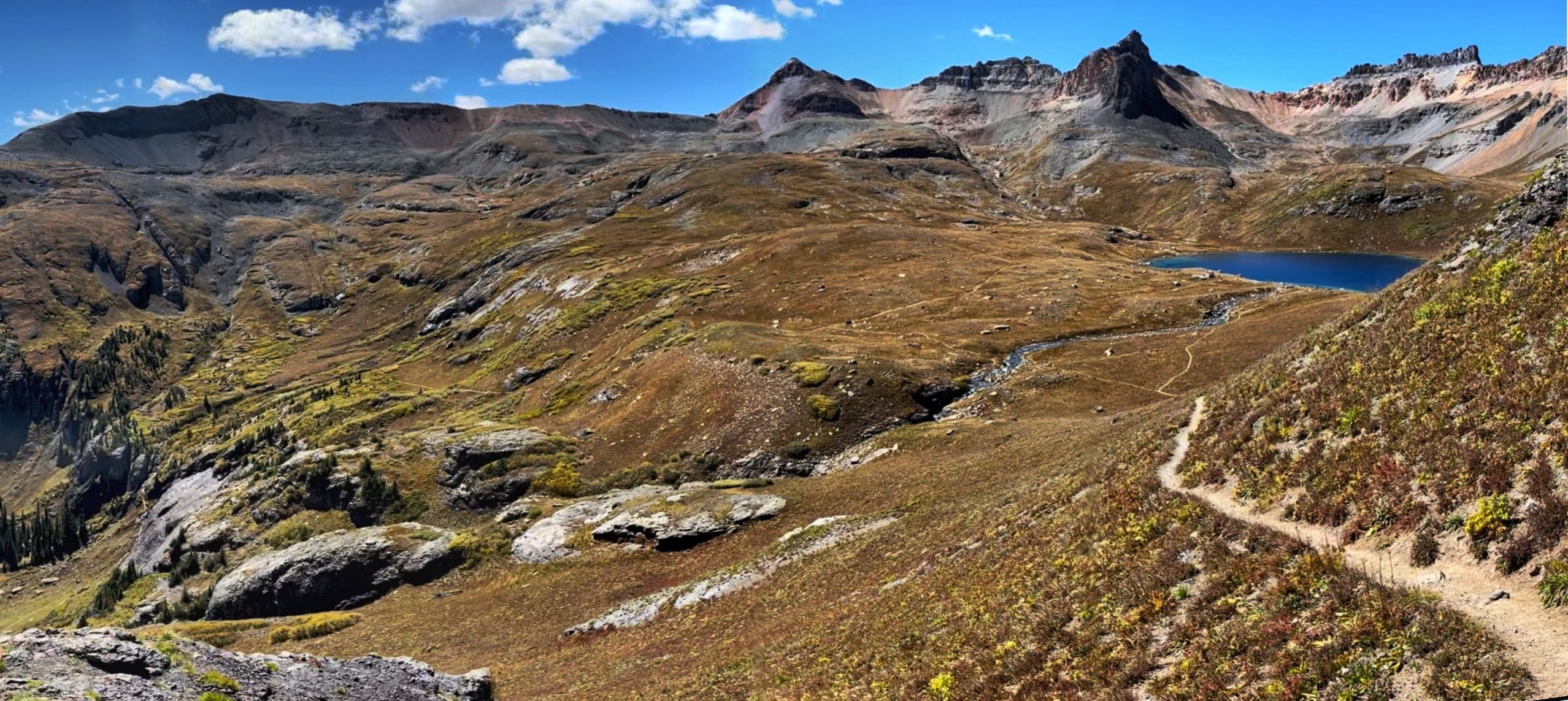 A panoramic view with a winding trail descending across a golden alpine tundra towards the blue waters of Ice Lake Colorado, set against a backdrop of jagged rocky peaks.


