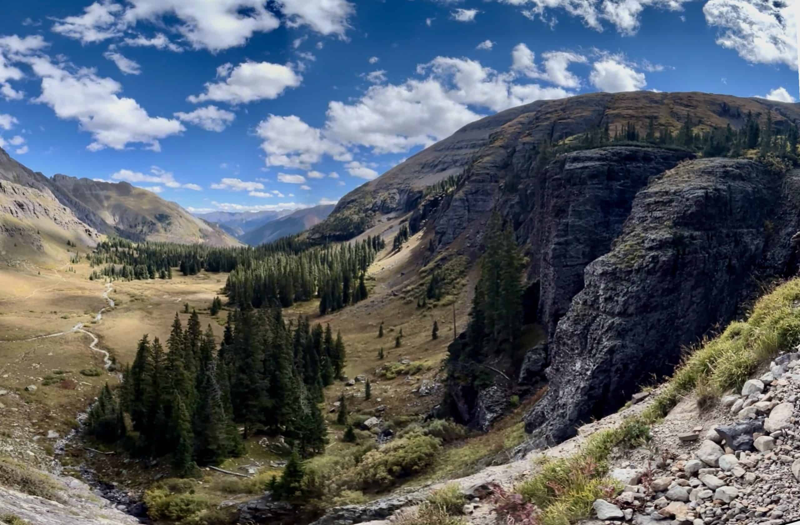 A rocky cliffside in the foreground overlooks a lush valley of evergreen trees and a winding blue stream descending toward a meadow below Ice Lake Colorado.
