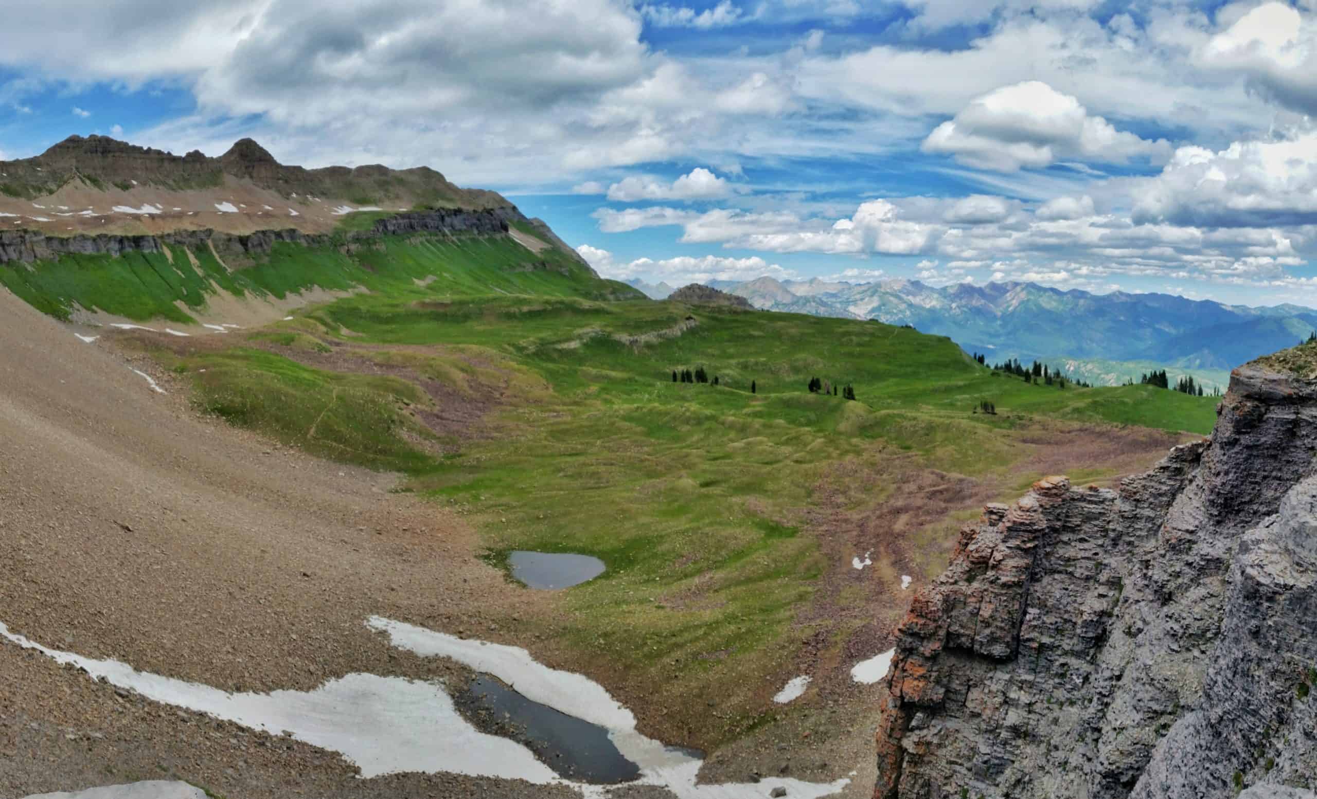 A beautiful open basin on the Mount Timpanogos hike, with a snowy rocky slope, a small pond and grassy meadows leading towards distant mountain peaks under a partly cloudy sky