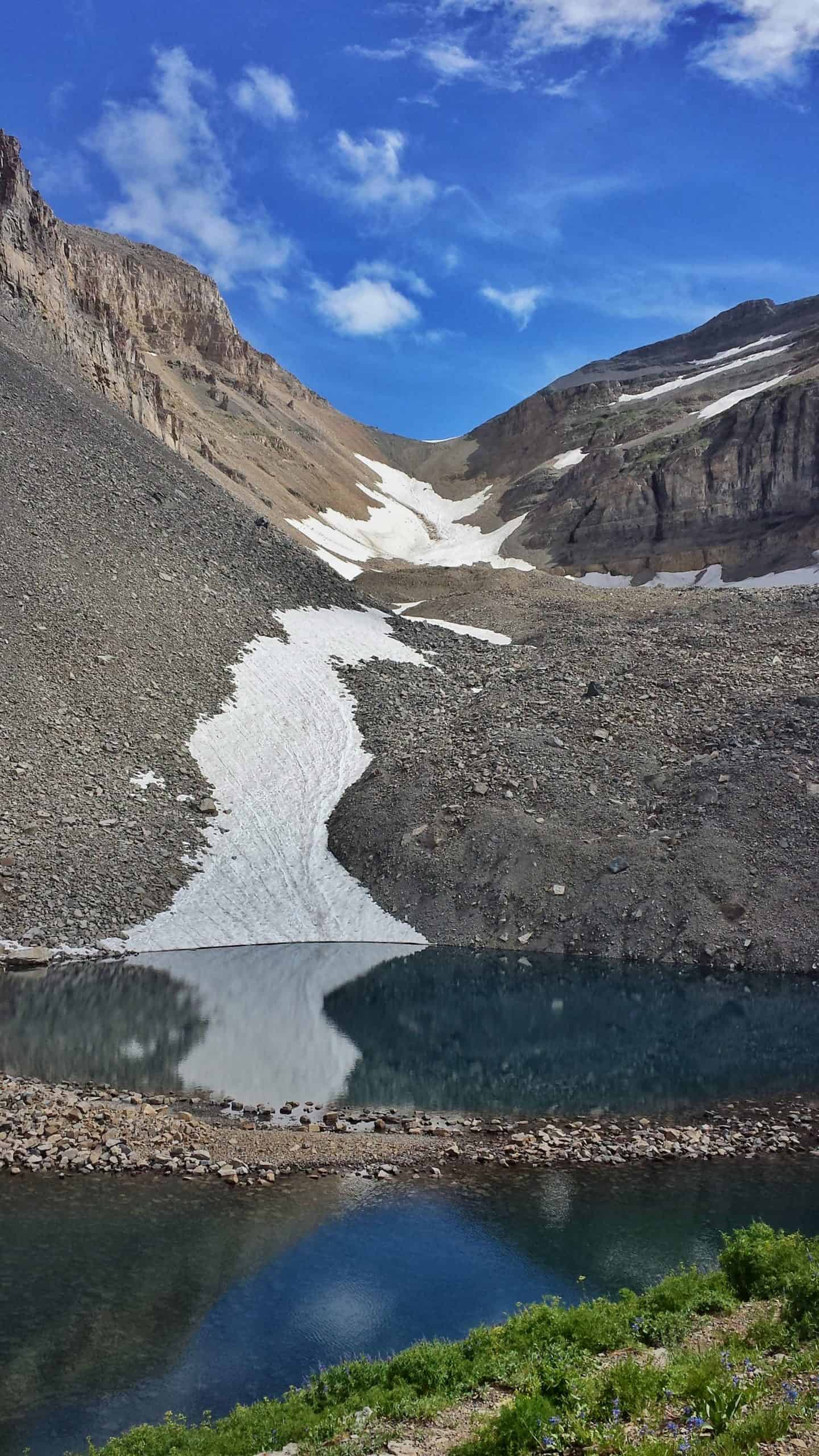 Along the Mount Timpanogos Hike, Emerald Lake mirrors the surrounding cliffs and blue sky, sitting at the foot of a steep, rocky slope marked by lingering snow patches.