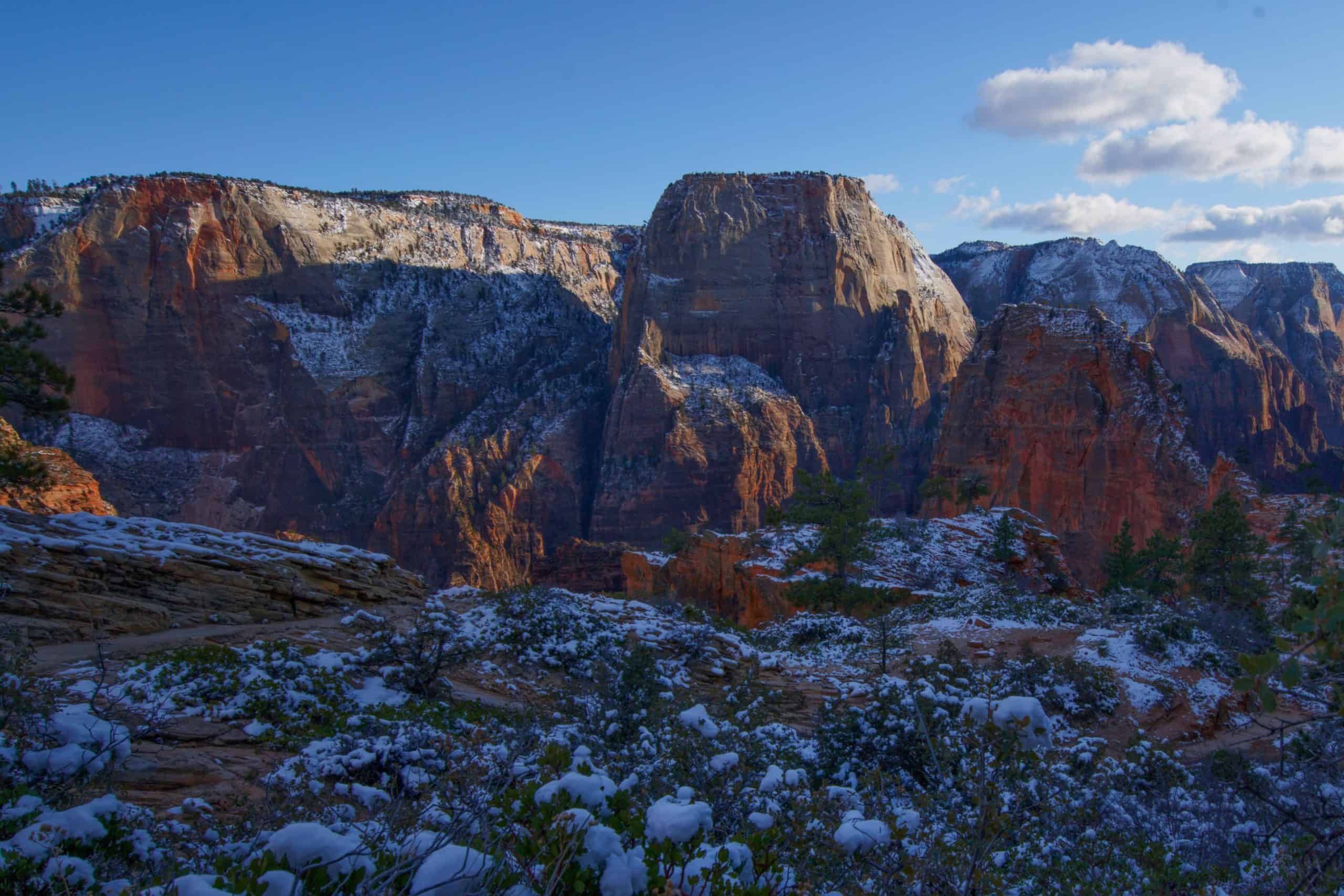 Snow-covered bushes and towering red rock cliffs dominate this view of Angels Landing, a popular destination for travelers following a 2 day Zion national park itinerary.

