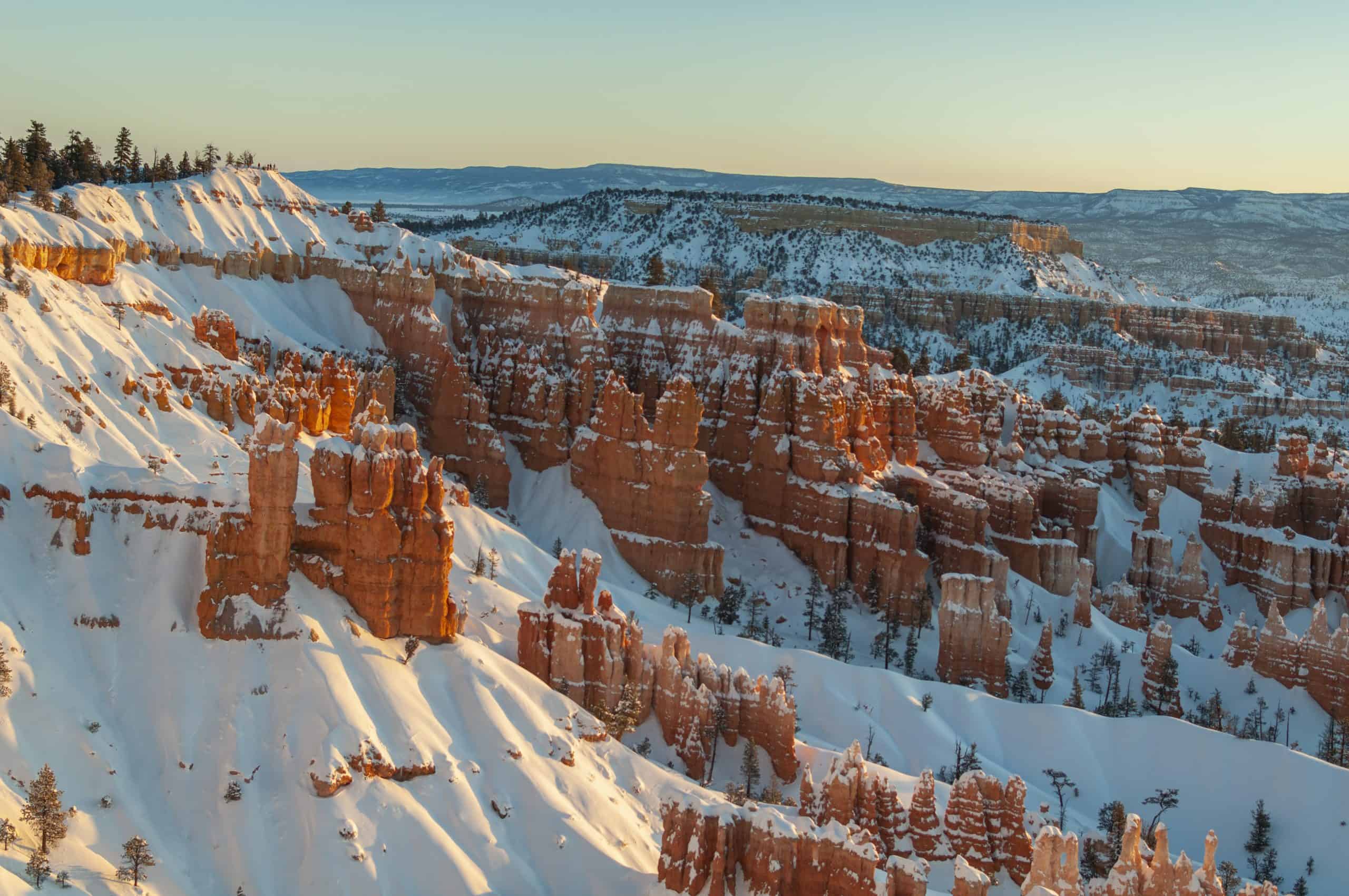A snowy winter scene of orange hoodoos in Bryce Canyon surrounded by wintry slopes under a light blue sky