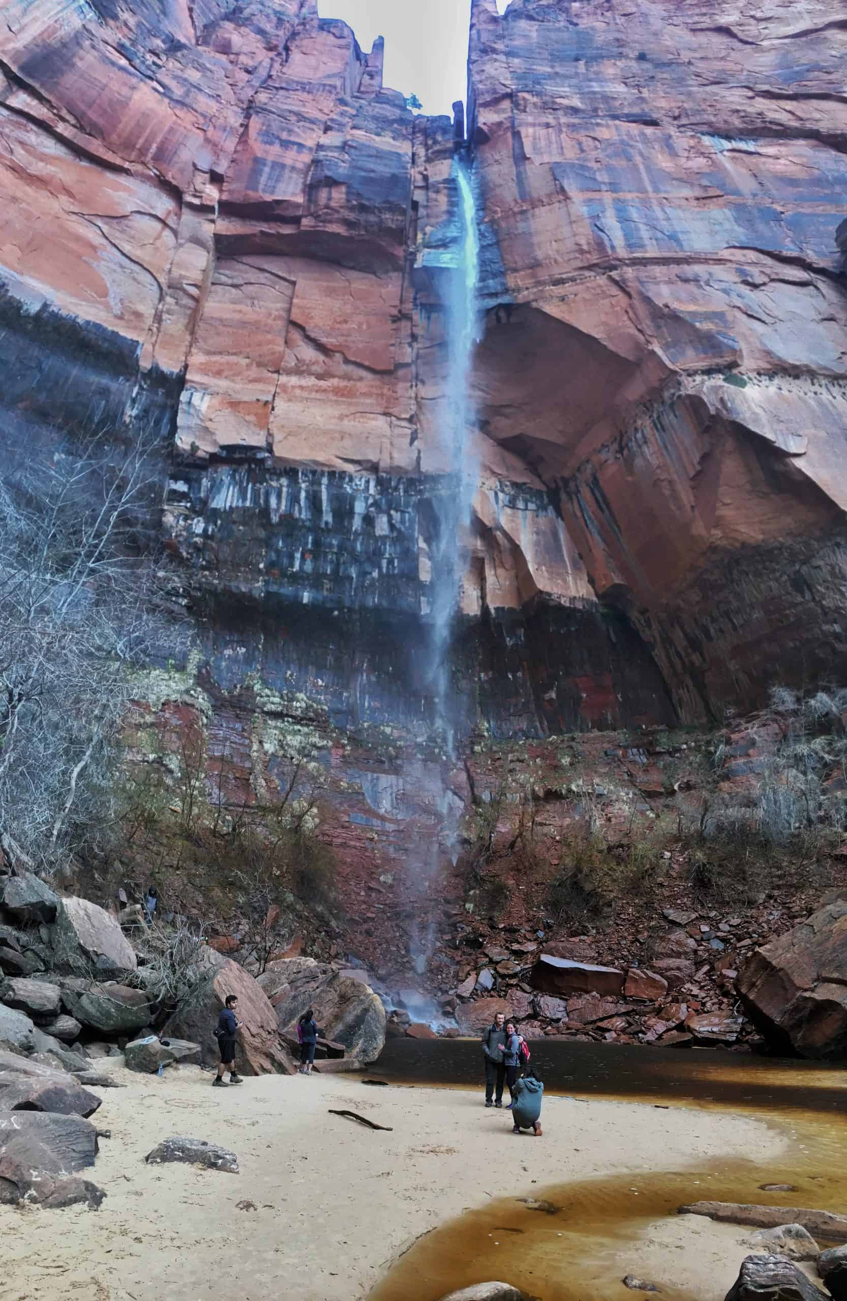 Hikers gather near the base of a tall waterfall plunging into the  Emerald Pools in Zion National Park.

